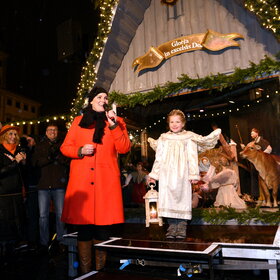 Vor dem Hintergrund einer lebensgroßen Weihnachtskrippe stehen eine Frau in rotem Mantel und ein als Engel gekleidetes blondes Kind, das eine Laterne hält.