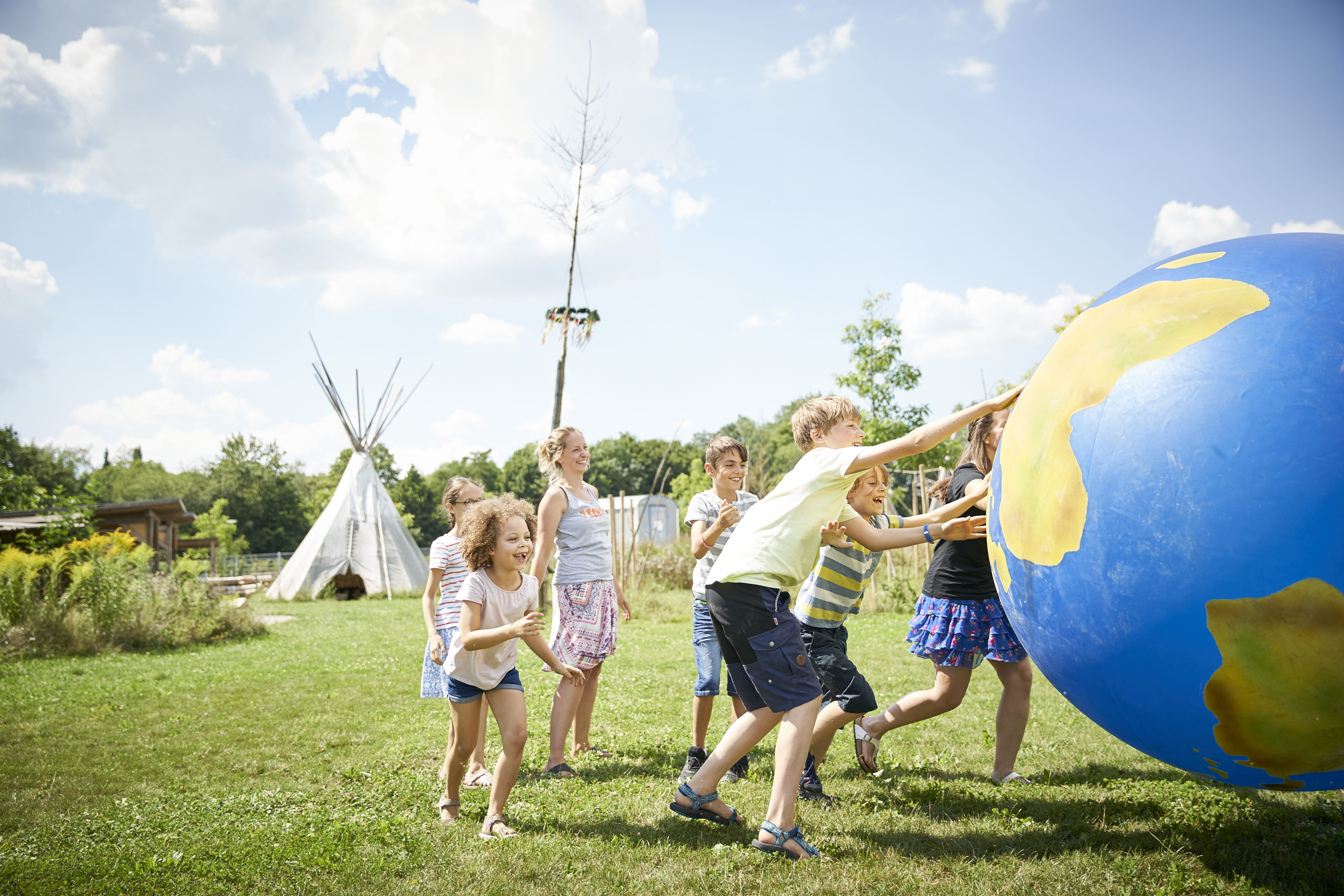 Sechs Kinder und Jugendliche rollen eine etwa zwei Meter große Weltkugel über eine Wiese. Im Hintergrund ist ein Indianer-Zelt zu sehen. 