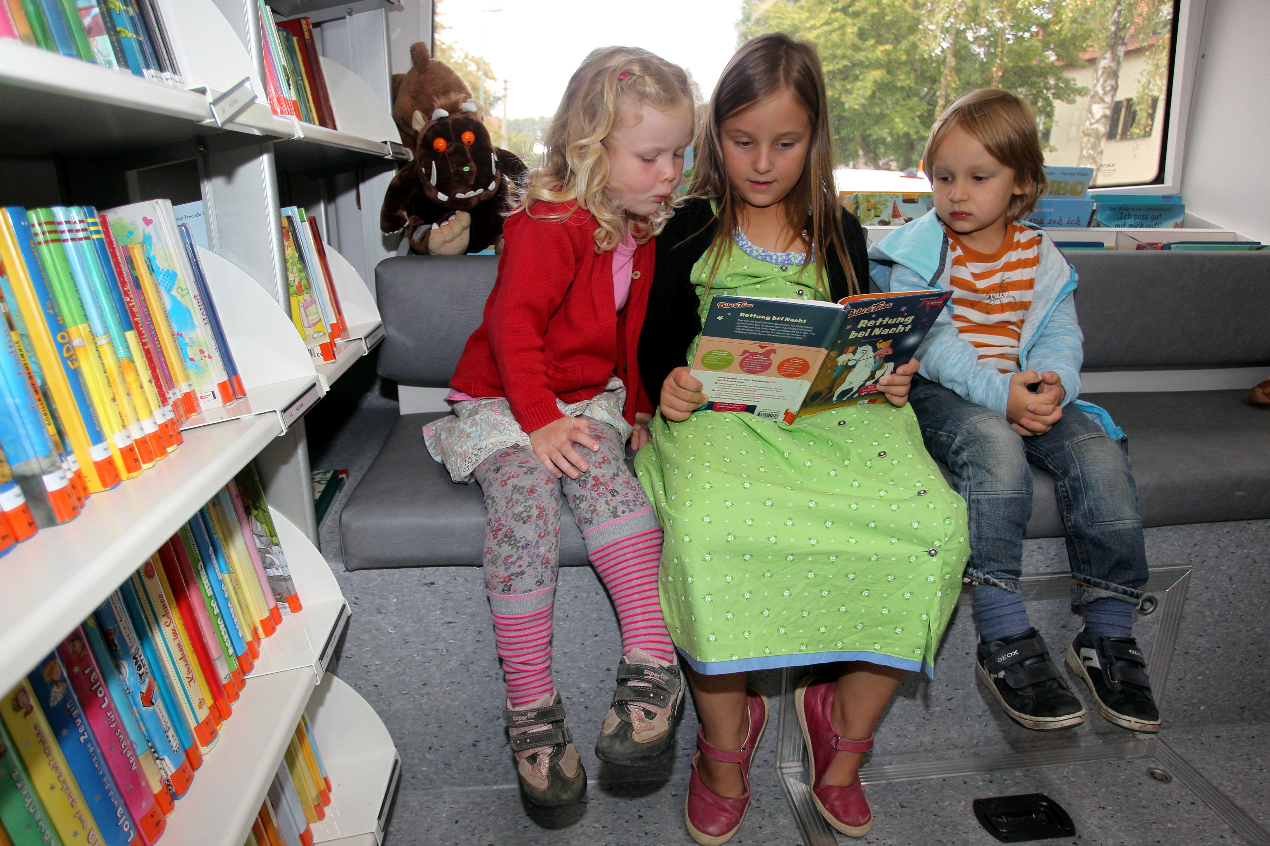 Drei Kinder sitzen auf dem Rücksitz in einem Bus. Neben ihnen ist ein Bücherregal zu sehen. Das Mädchen in der Mitte hat ein aufgeschlagenes Buch auf dem Schoß. Das kleinere Mädchen links und der kleine Junge rechts schauen ins Buch.
