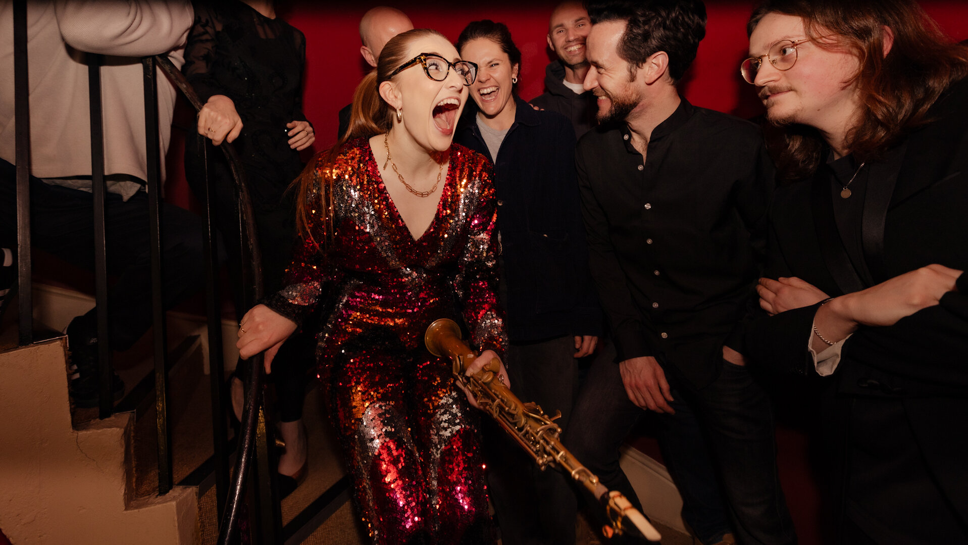 Eine Frau in einem Glitzerkleid und mit einem Saxophon in der Hand lacht laut. Um sie herum stehen lachende Personen in schwarzer Kleidung.
