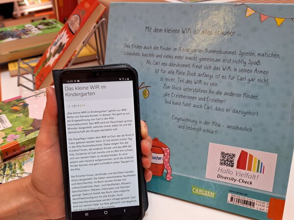 Die Rückseite des Bilderbuchs, auf der der Aufkleber des Projekts mit QR-Code klebt. Eine Hand hält ein Smartphone mit dem Bewertungstext zum Buch hoch.
