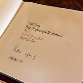 Es ist ein geöffnetes Buch mit Text und Unterschrift zu sehen.
