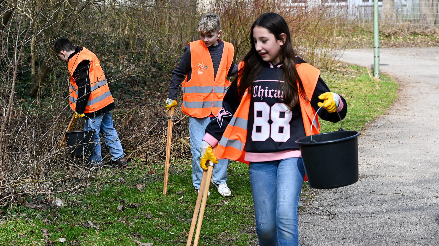 Das Bild zeigt Kinder in orangenen Warnwesten, ausgestattet mit Holzzangen, gelben Handschuen und schwarzen Eimern in der Hand. Sie laufen an einer Wiese entlang und heben  Müll auf