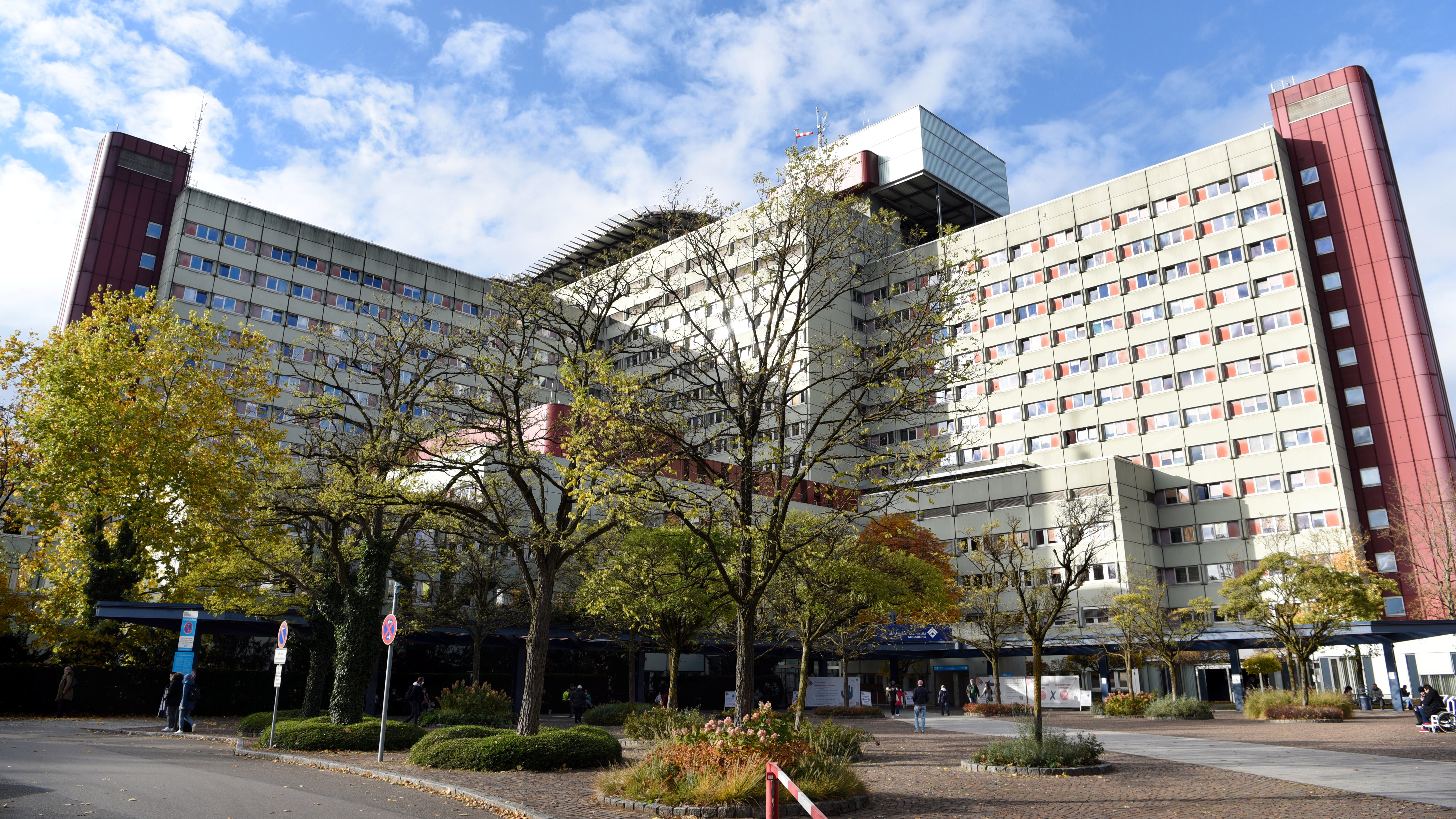 Das Foto zeigt ein weißes, hohes Gebäude, das eckig ist. Im Vordergrund sind Bäume zu sehen