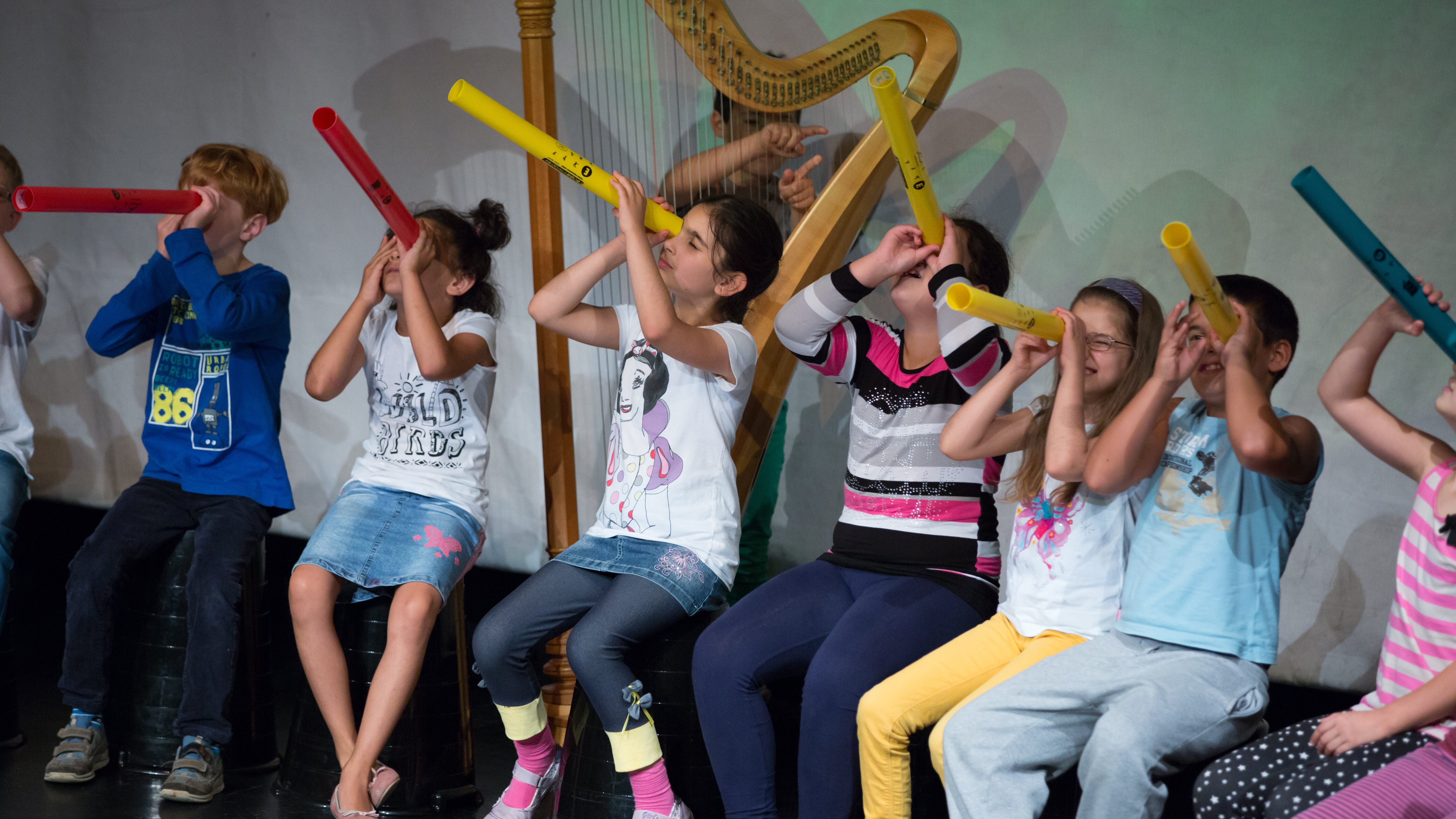 Das Foto zeigt Kinder, die auf einer Bühne sitzen und Boomwhackers in der Hand halten. Das sind lange bunte Plastikröhren, die je nach länge einen unterschiedlichen Ton machen, wenn man sie z.B. gegen eine Hand schlägt.