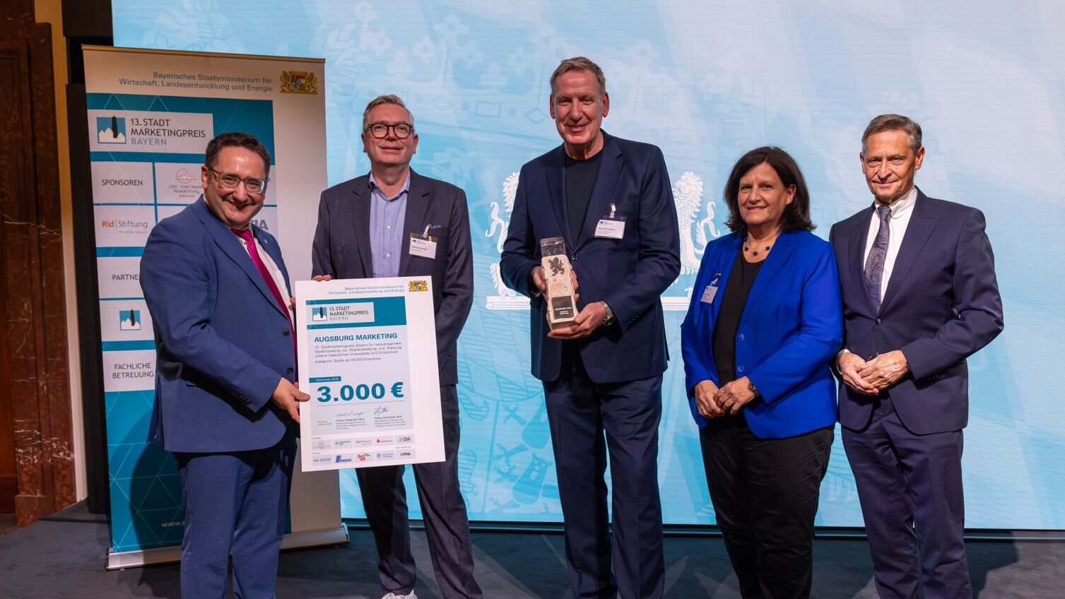 5 Personen stehen nebeneinander, eine hält eine Tafel mit der Gewinnerurkunde