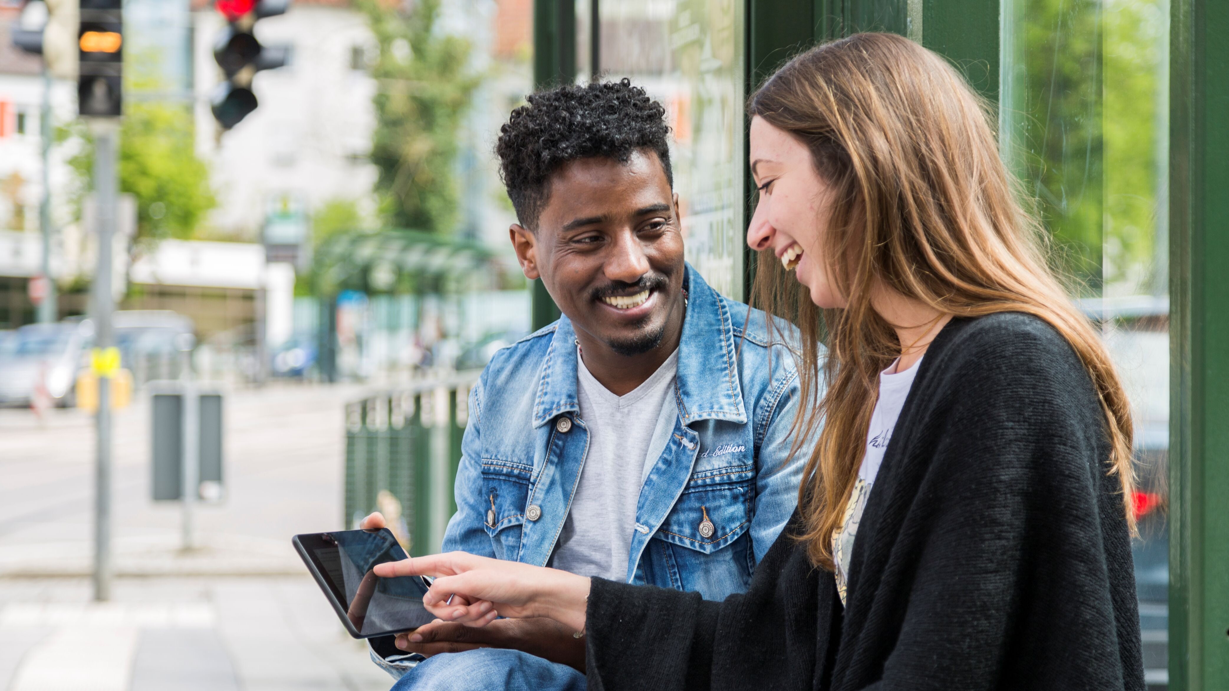zwei jüngere Menschen - ein dunkelhäutiger Mann und eine eurasische Frau - sitzen an einer Haltetselle und sprechen miteinander, die Frau hält ein Handy