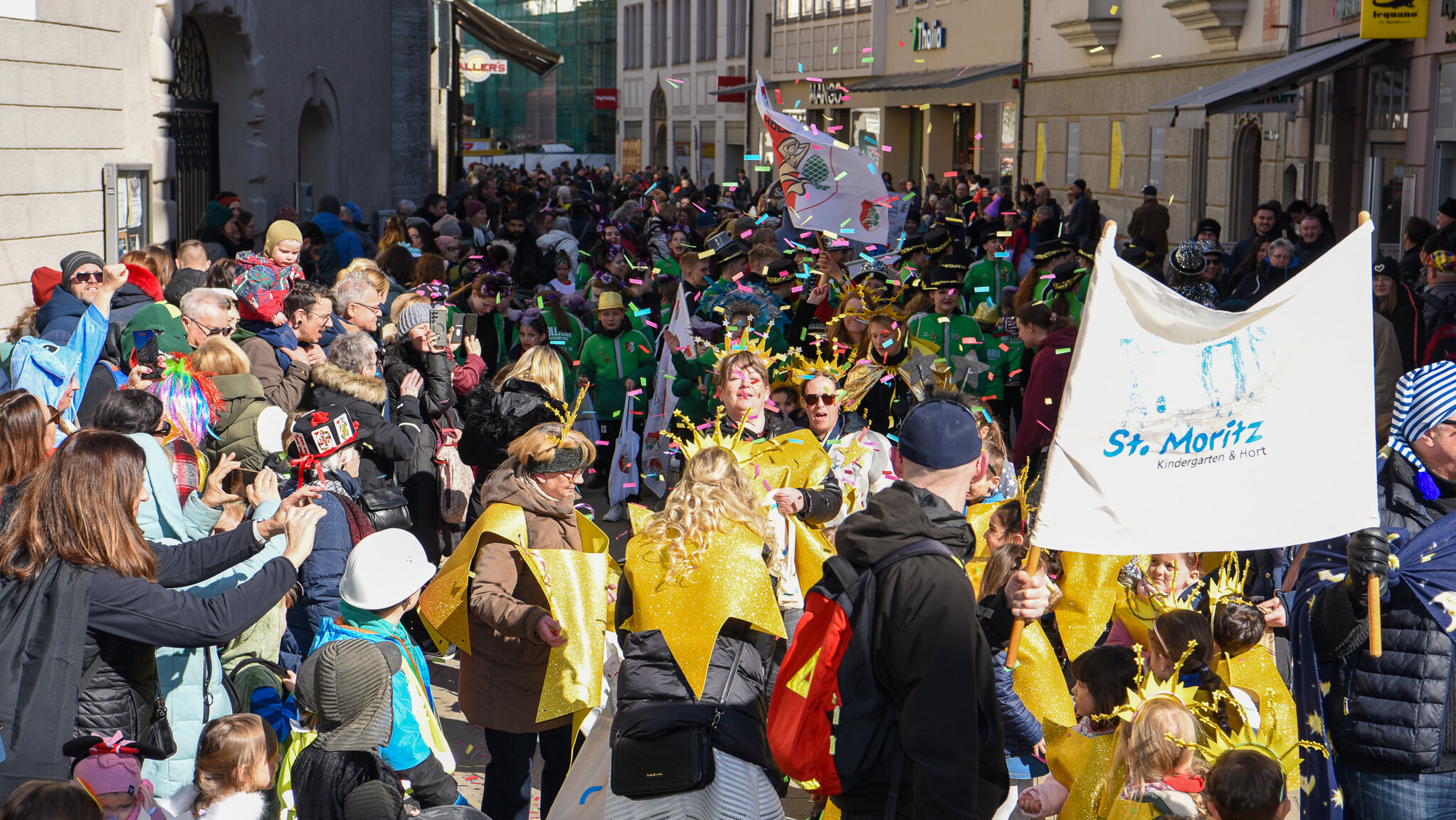 Zum Fasching verkleidete Menschen in einer Straße. In der Mitte des Bilds wird Konfetti in die Luft geworfen