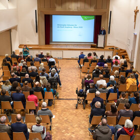 Blick von der Empore in einen vollbesetzten Saal; an der Stirnseite eine große Projektionsfläche, darauf die Worte "Aktionsplan Inklusion der Stadt Augsburg - Bilanz 2025"