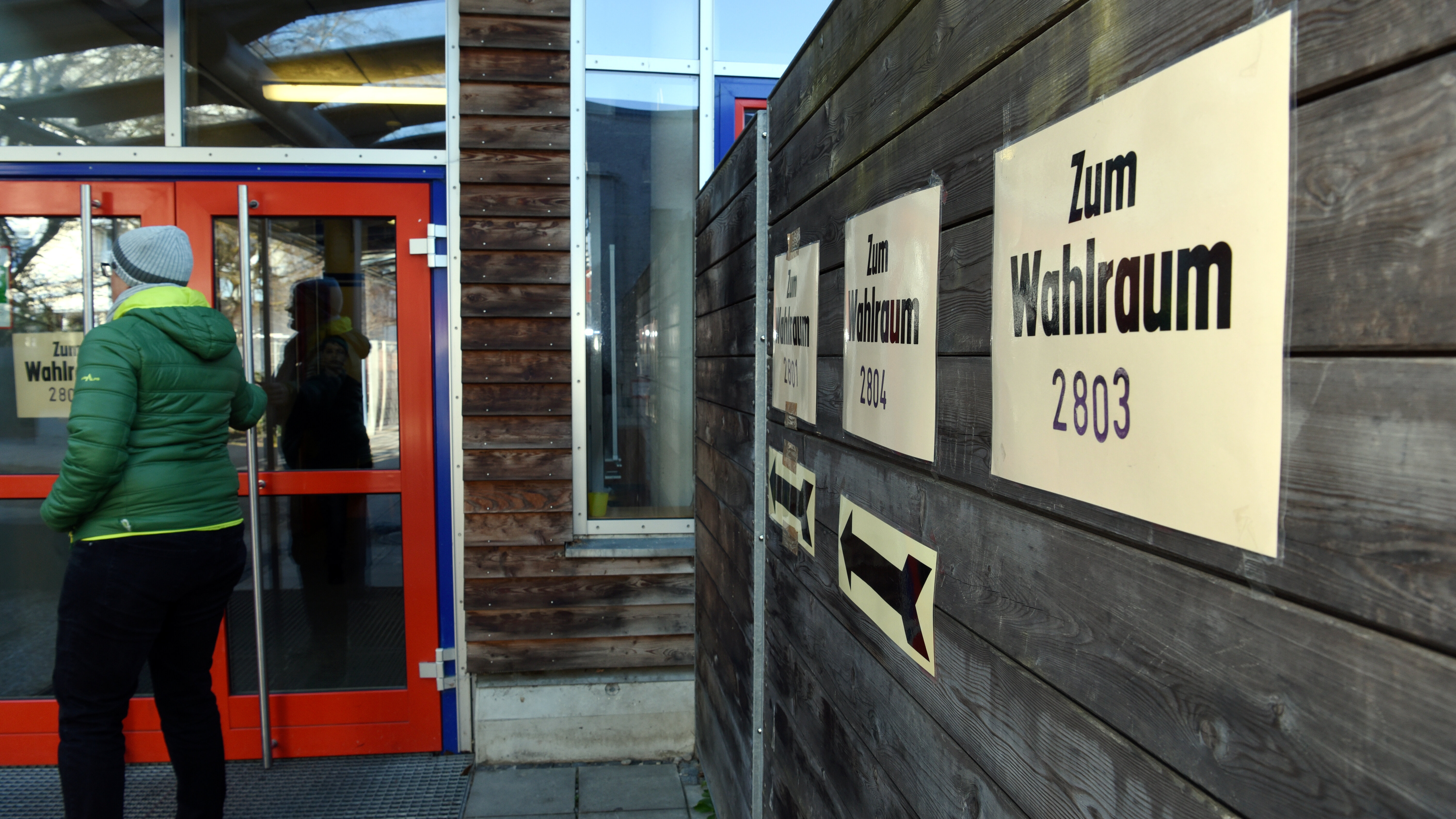 Ein Gebäude, jemand geht durch die Türe. An der Wand hängt ein Schild "Zum Wahlraum"