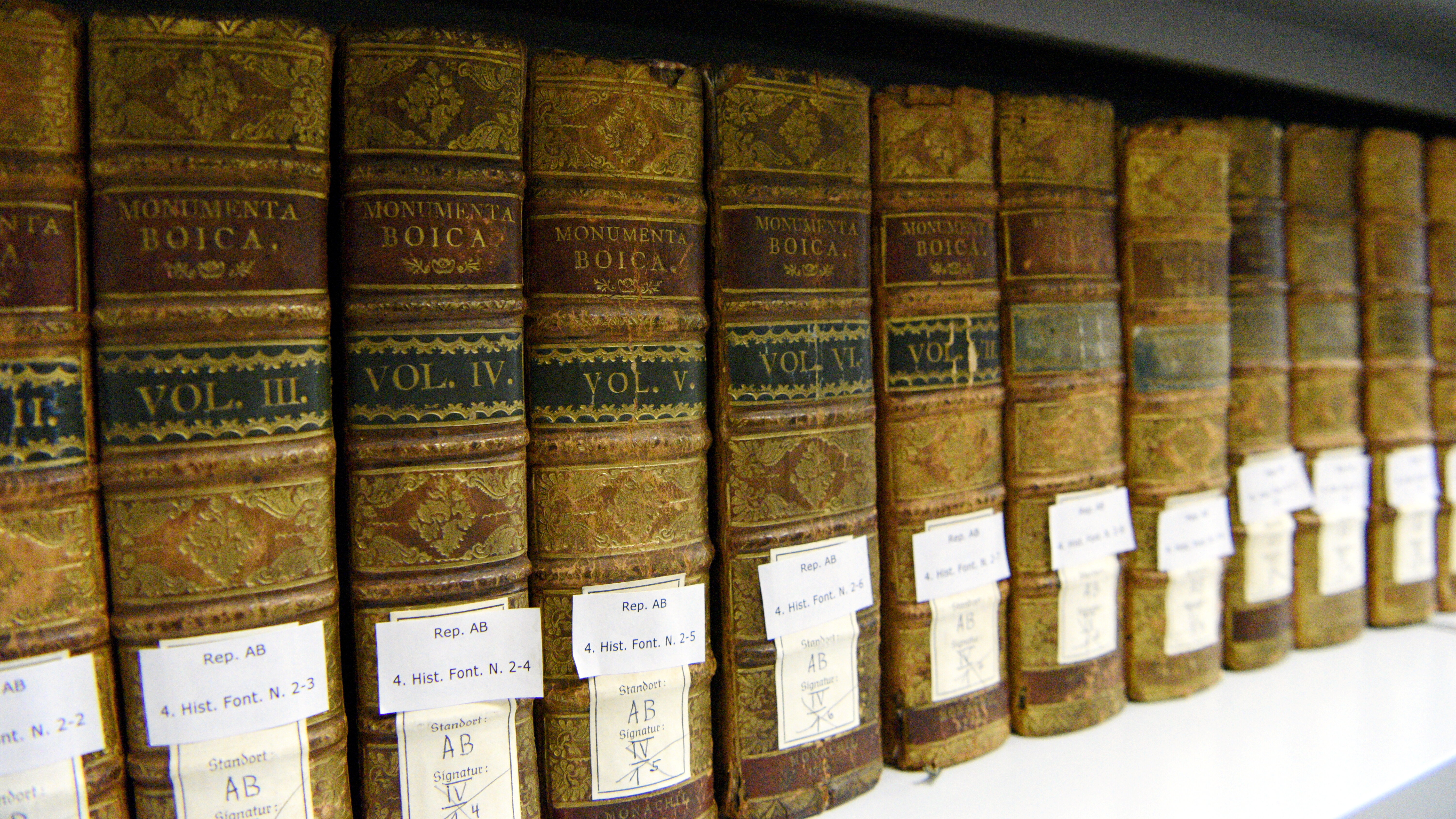 Das Foto zeigt alte, reich verzierte Bücher, die in einem Regal stehen. Am Buchrücken sind kleine Zettelchen mit Nummern und Buchstaben angebracht