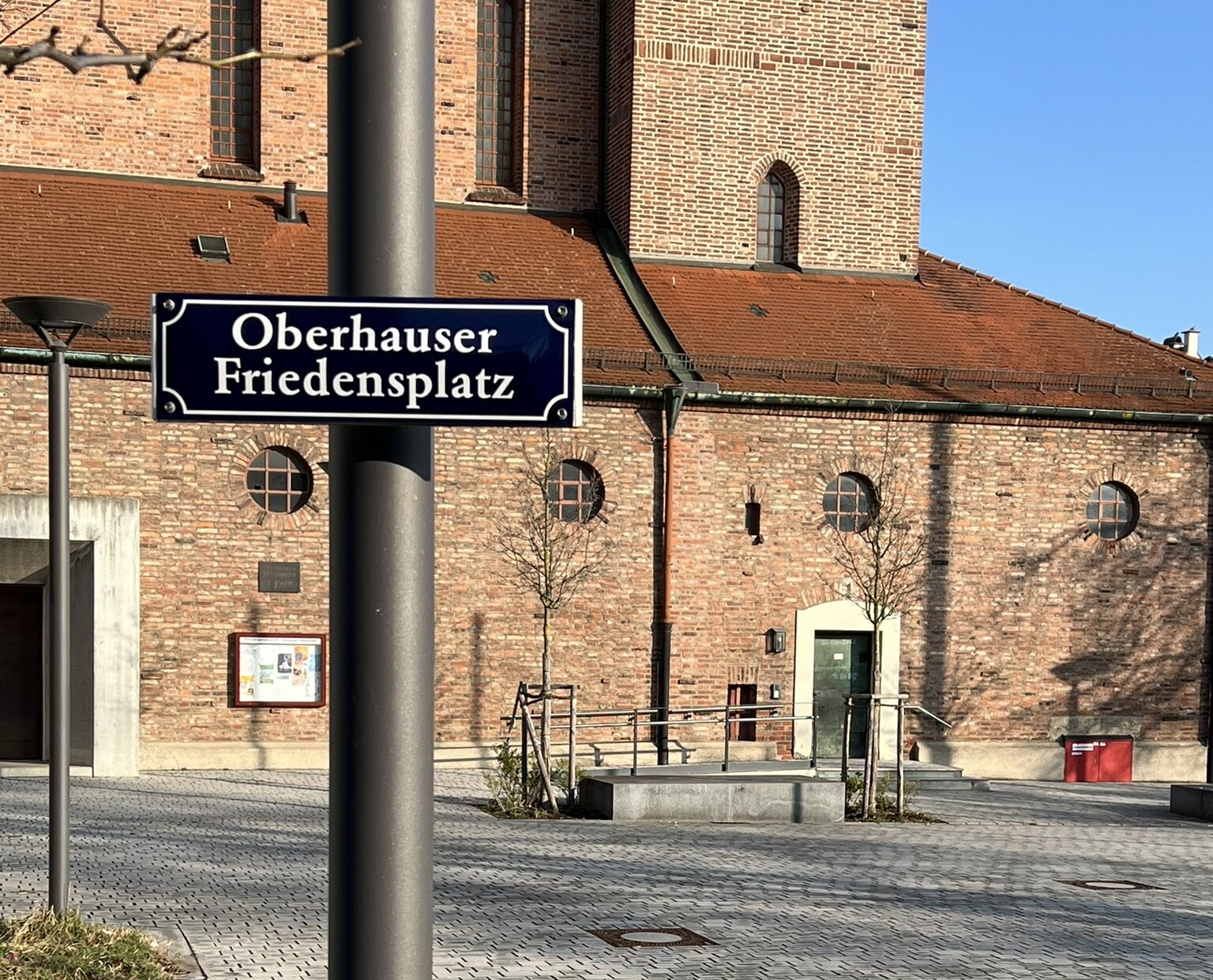 Blick auf einen Platz mit Straßenschild "Oberhauser Friedensplatz", im Hintergrund eine Backsteinkirche