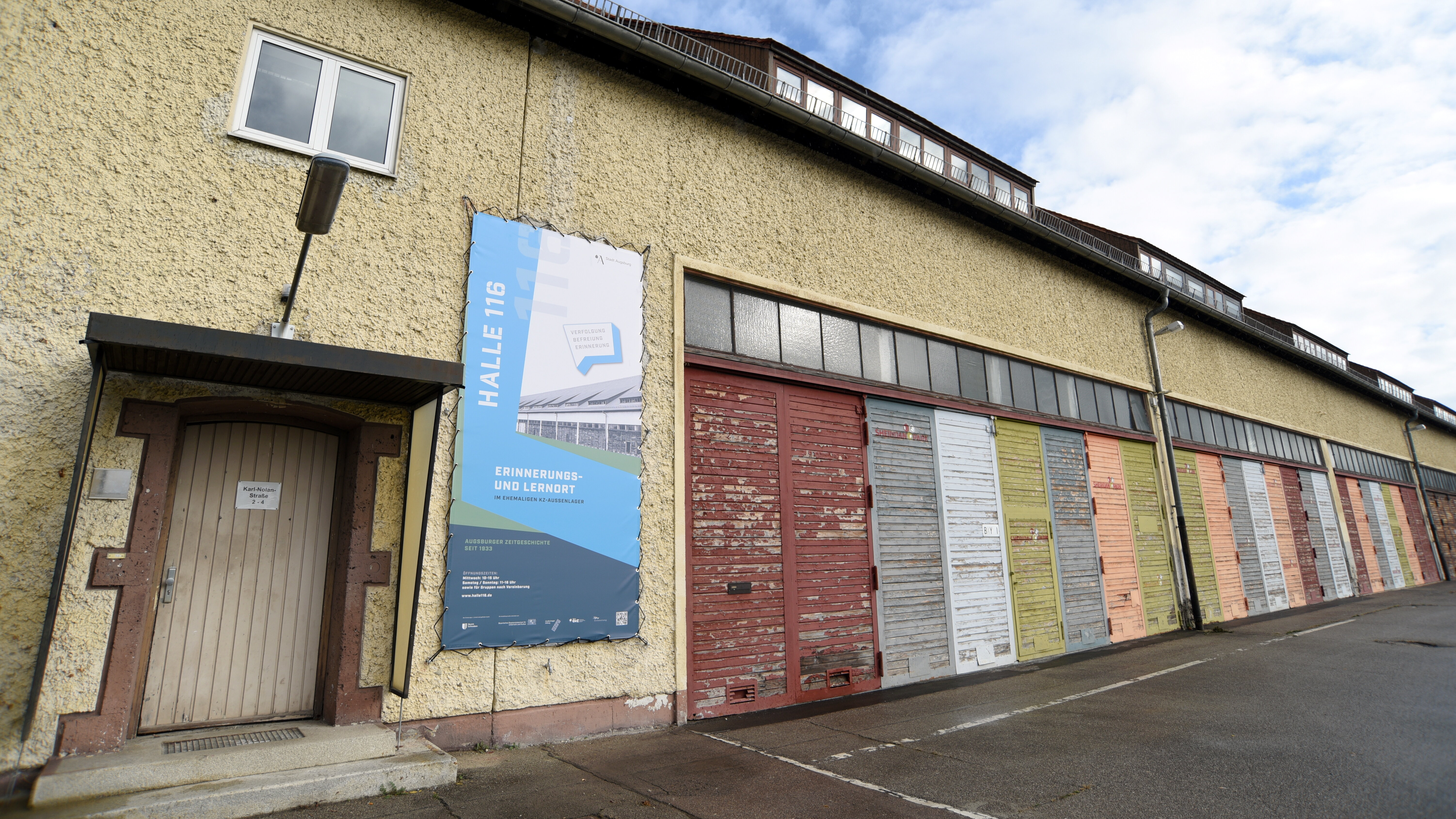 Lagerhalle mit Schiebetüren in verschiedenen Farben; neben dem Eingang ein großes Plakat, das auf den Erinnerungs- und Lernort hinweist