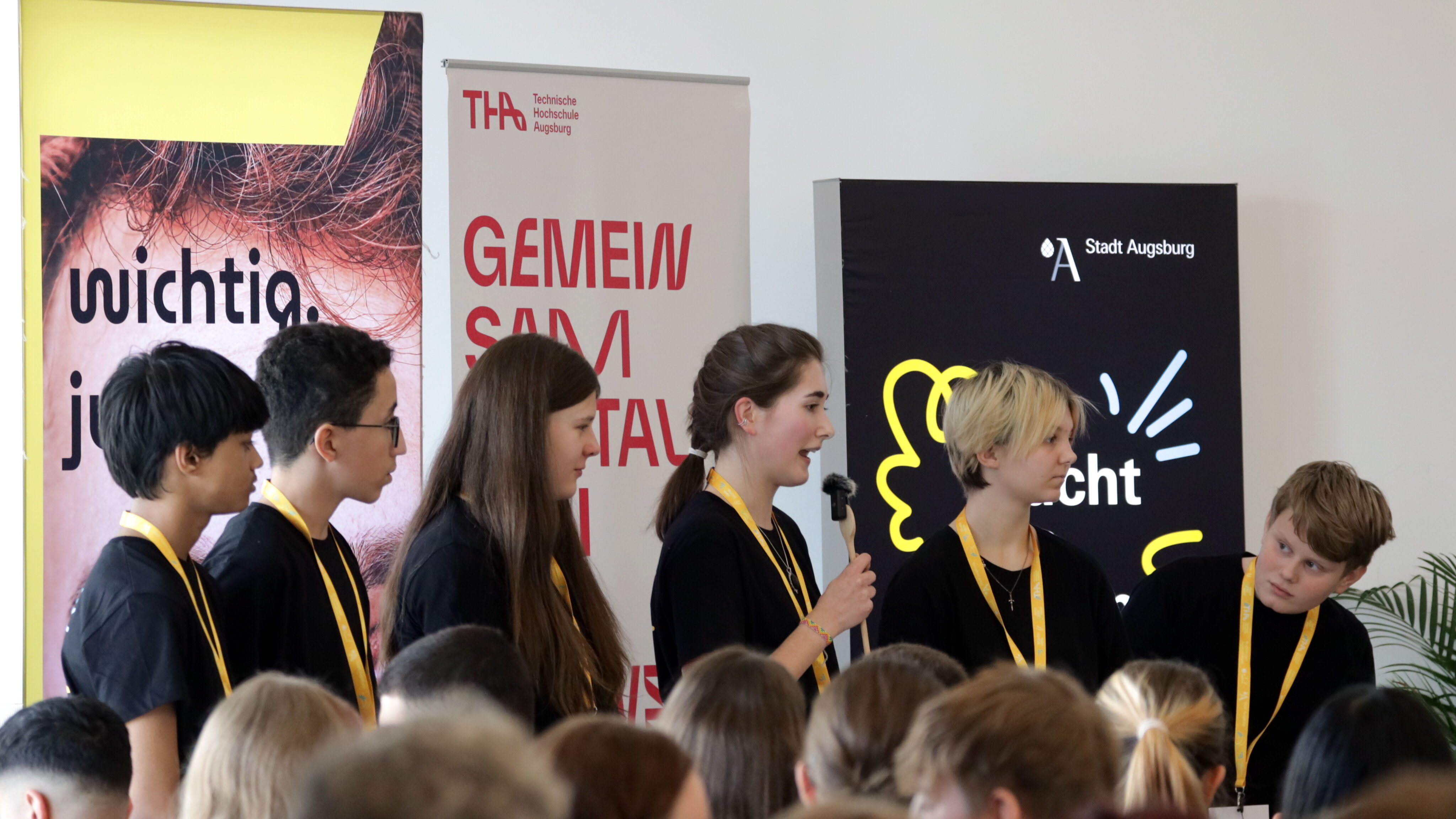 drei Mädchen und drei Jungen auf einer Bühne vor Roll-ups, ein Mädchen spricht in ein Mikrofon