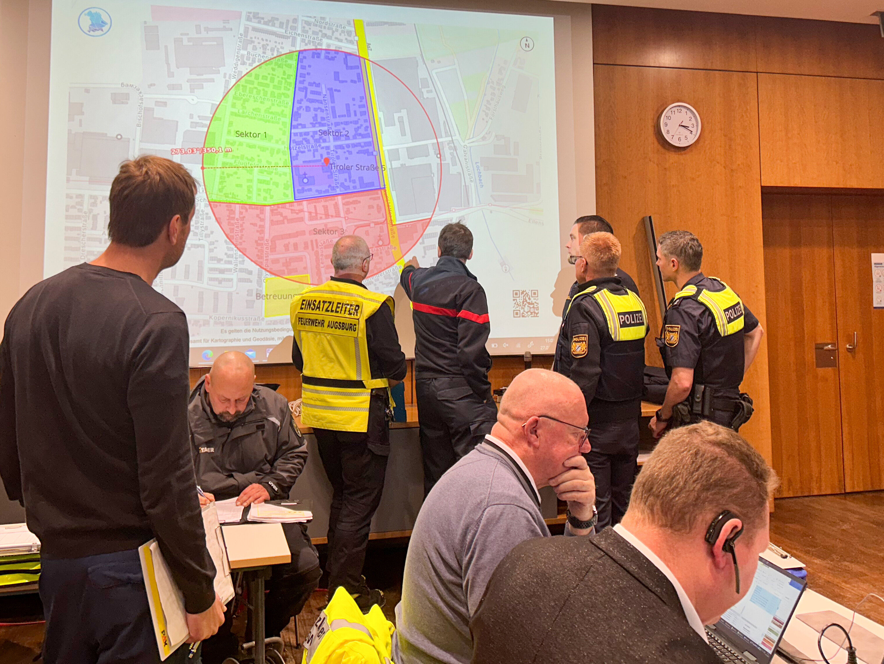 Einsatzkräfte von Polizei und Feuerwehr betrachten eine eingezeichnete Sperrzone auf einer Großleinwand. 