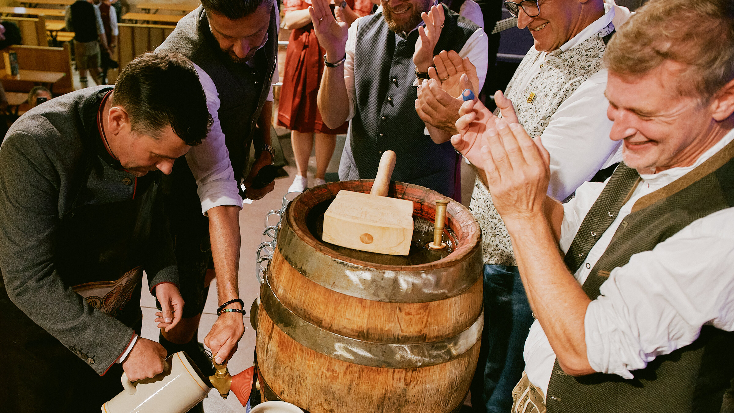 Augsburgs Oberbürgermeister Dr Florian Freund steht vor einem Bierfaß und zapft die ersten Biere zur Eröffnung des Plärrers. Er träft eine Lederschürze. Umstehende Personen applaudieren. 