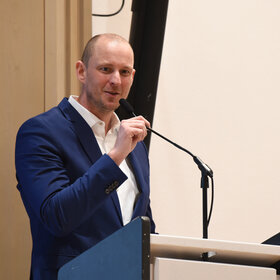 Mann mit Stirnglatze und Anzug am Rednerpult, eine Hand am Mikrofon