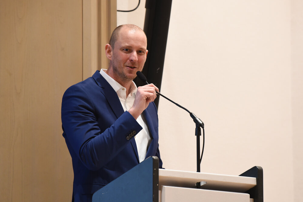 Mann mit Stirnglatze und Anzug am Rednerpult, eine Hand am Mikrofon