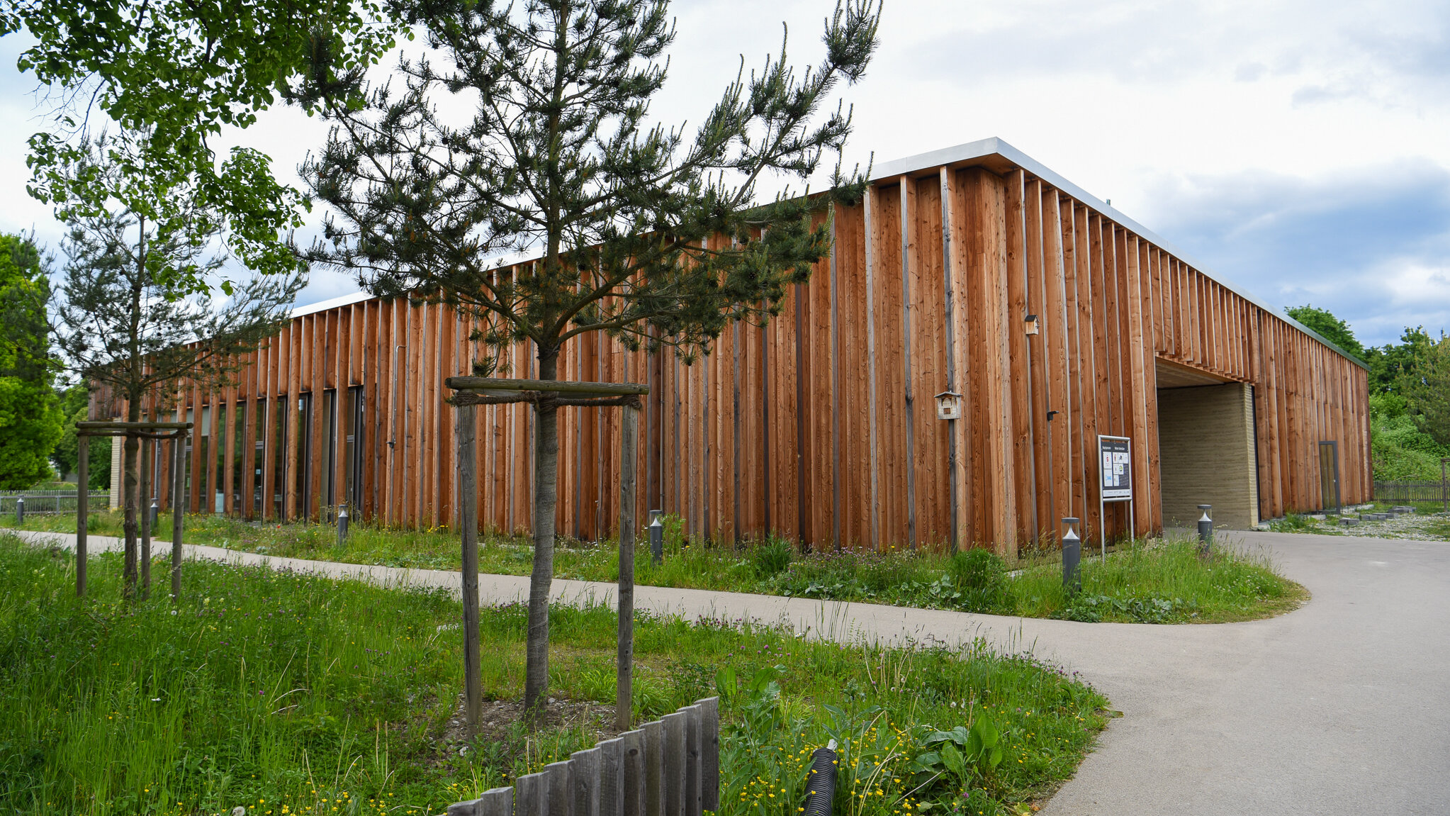 holzverkleidetes Gebäude von außen, im Vordergrund Wiese und ein Baum