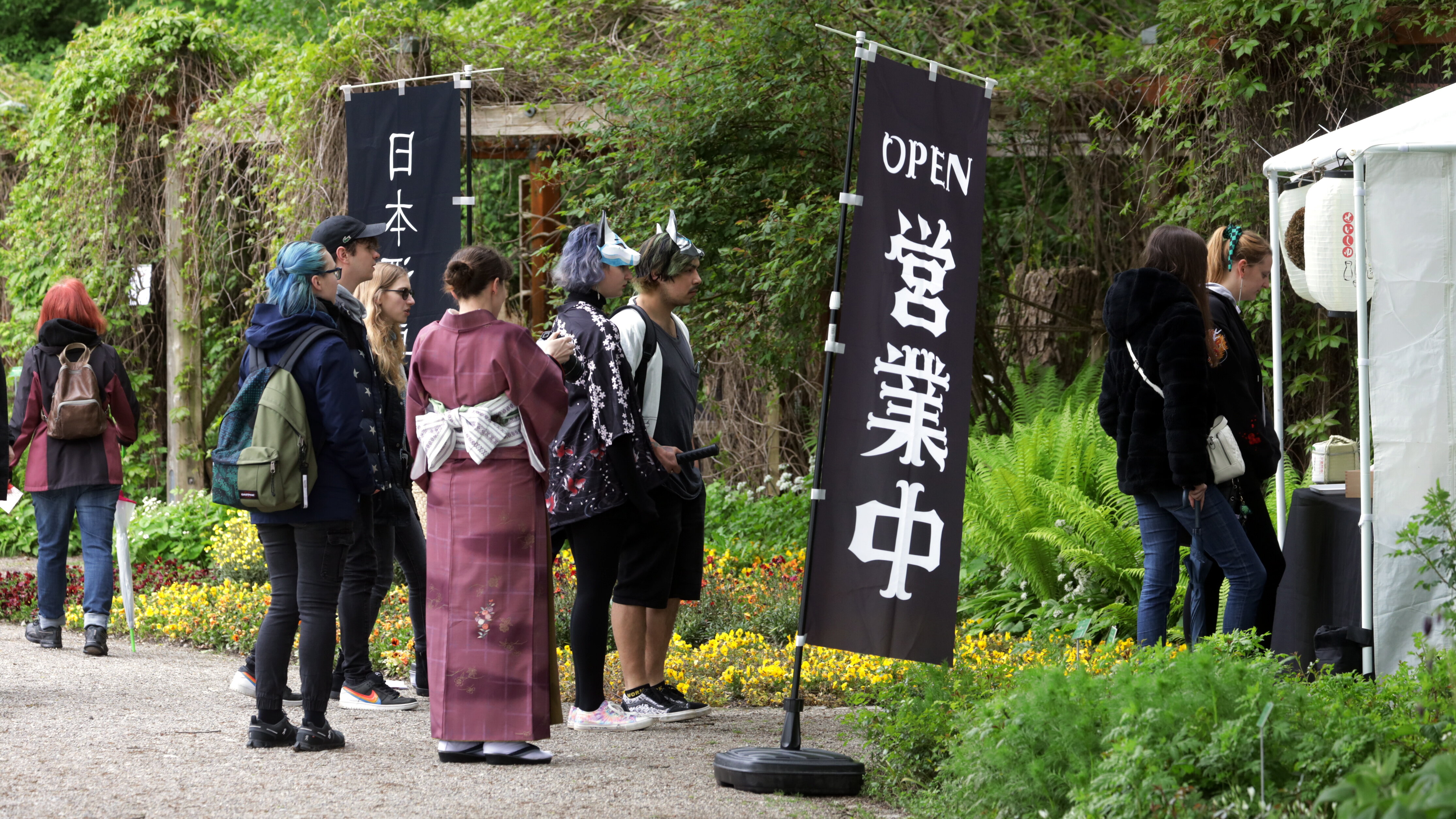 Menschen, darunter auch eine Frau im Kimono, stehen vor einem Zeltpavillon, vor dem 2 Hinweisfahnen mit japanischen Schriftzeichen stehen