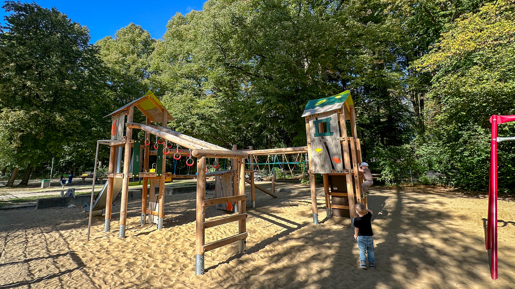 Ein Klettergerät aus Holz auf einem Spielplatz. Im Hintergrund sind hohe Bäume. Zwei Kinder spielen am Gerät