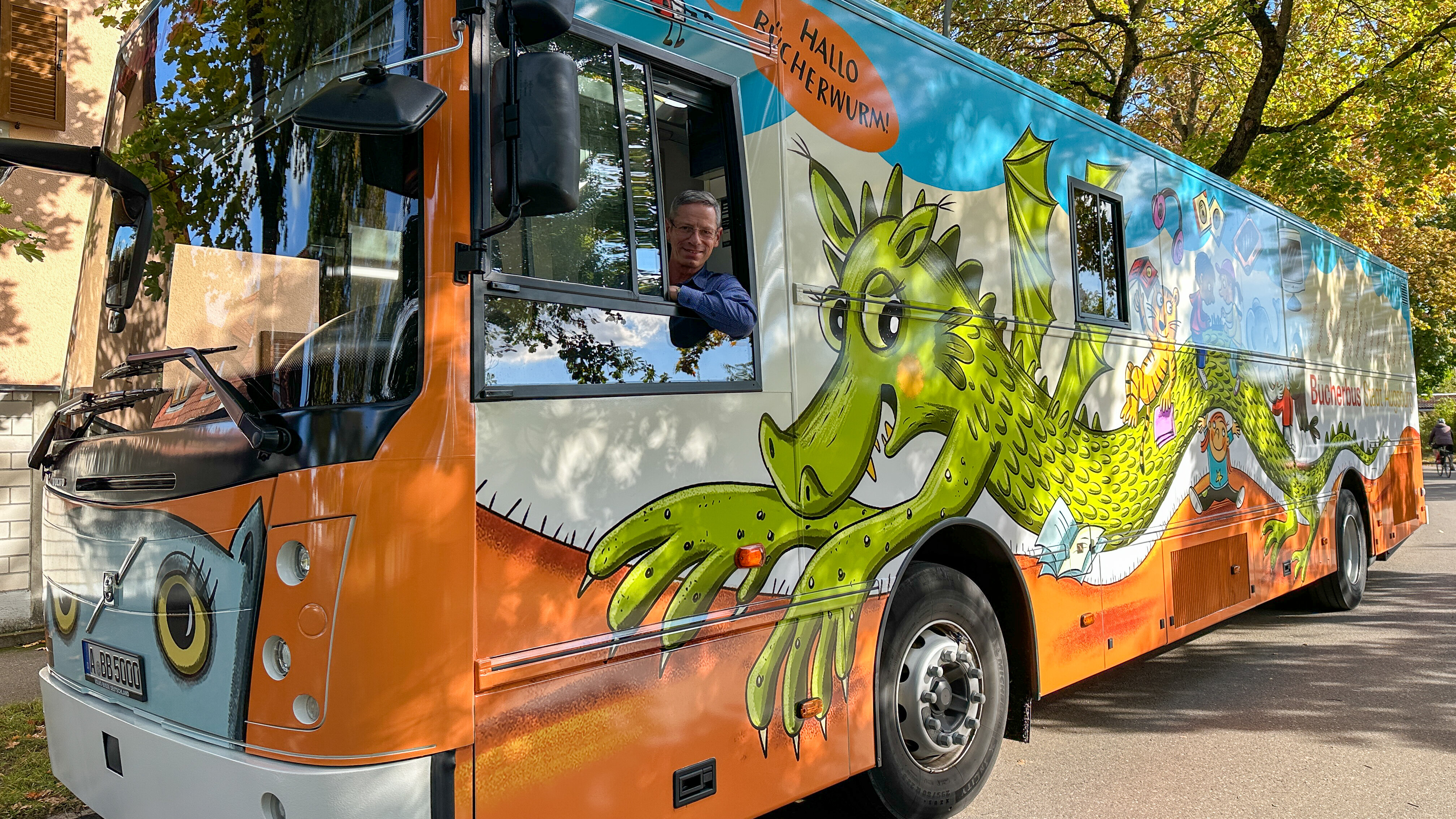 Das Bild zeigt den Bücherbus. Auf der Seite ist ein grüner Drache, das restliche Design ist in blau und orange gehalten