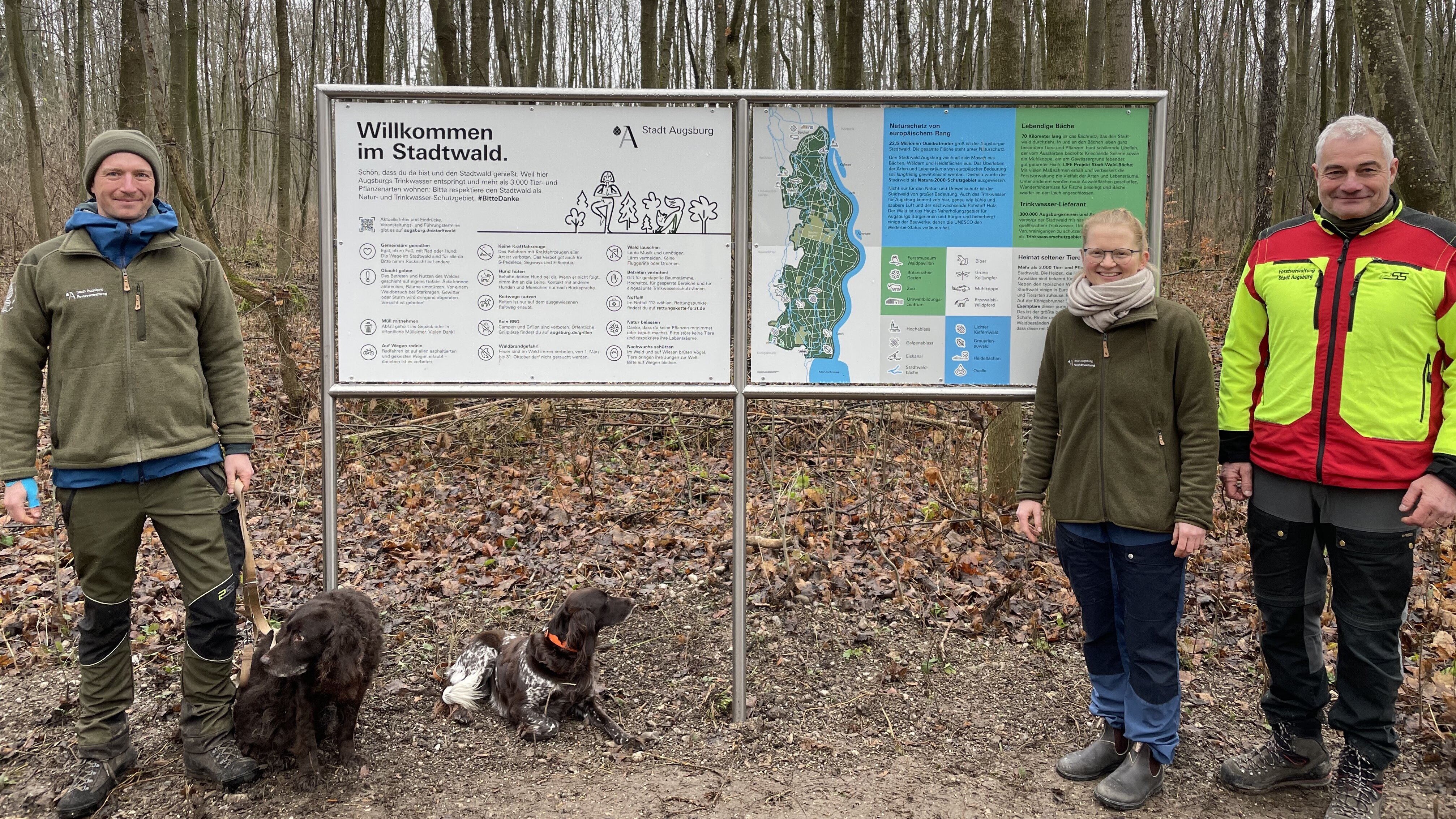 Drei Menschen in Arbeitskleidung stehen vor Infotafeln. Auf einer steht groß Willkommen im Stadtwald. Im Hintergrund ist ein herbstlicher Wald mit kahlen Bäumen zu sehen