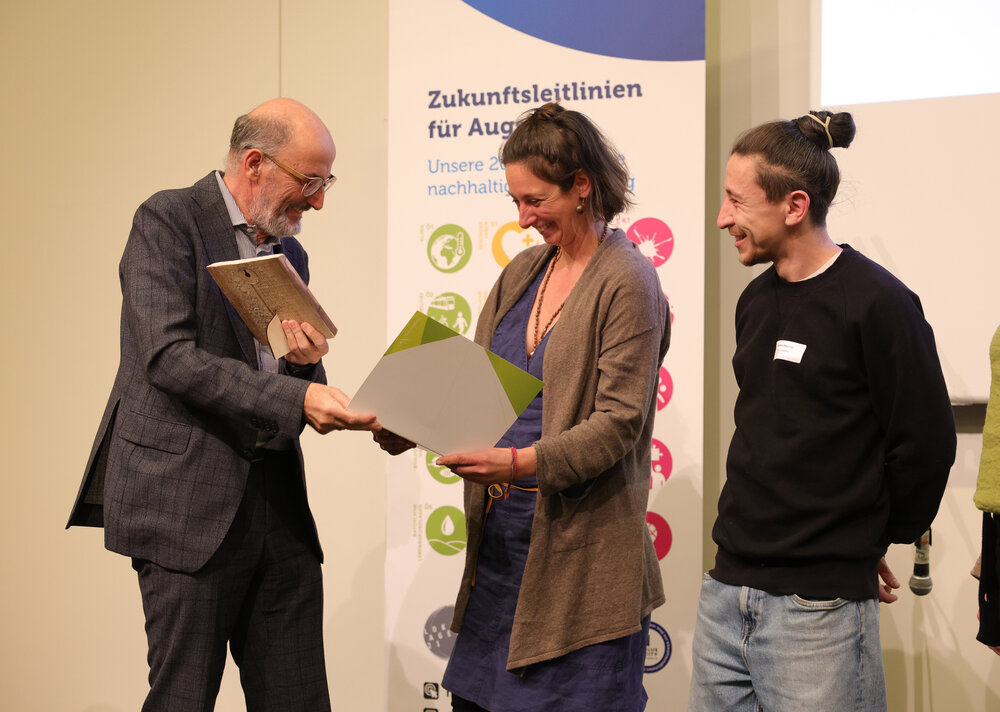 Mann im Anzug überreicht Urkunde an eine Frau, ein junger Mann mit Dutt steht daneben