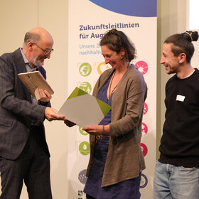 Mann im Anzug überreicht Urkunde an eine Frau, ein junger Mann mit Dutt steht daneben