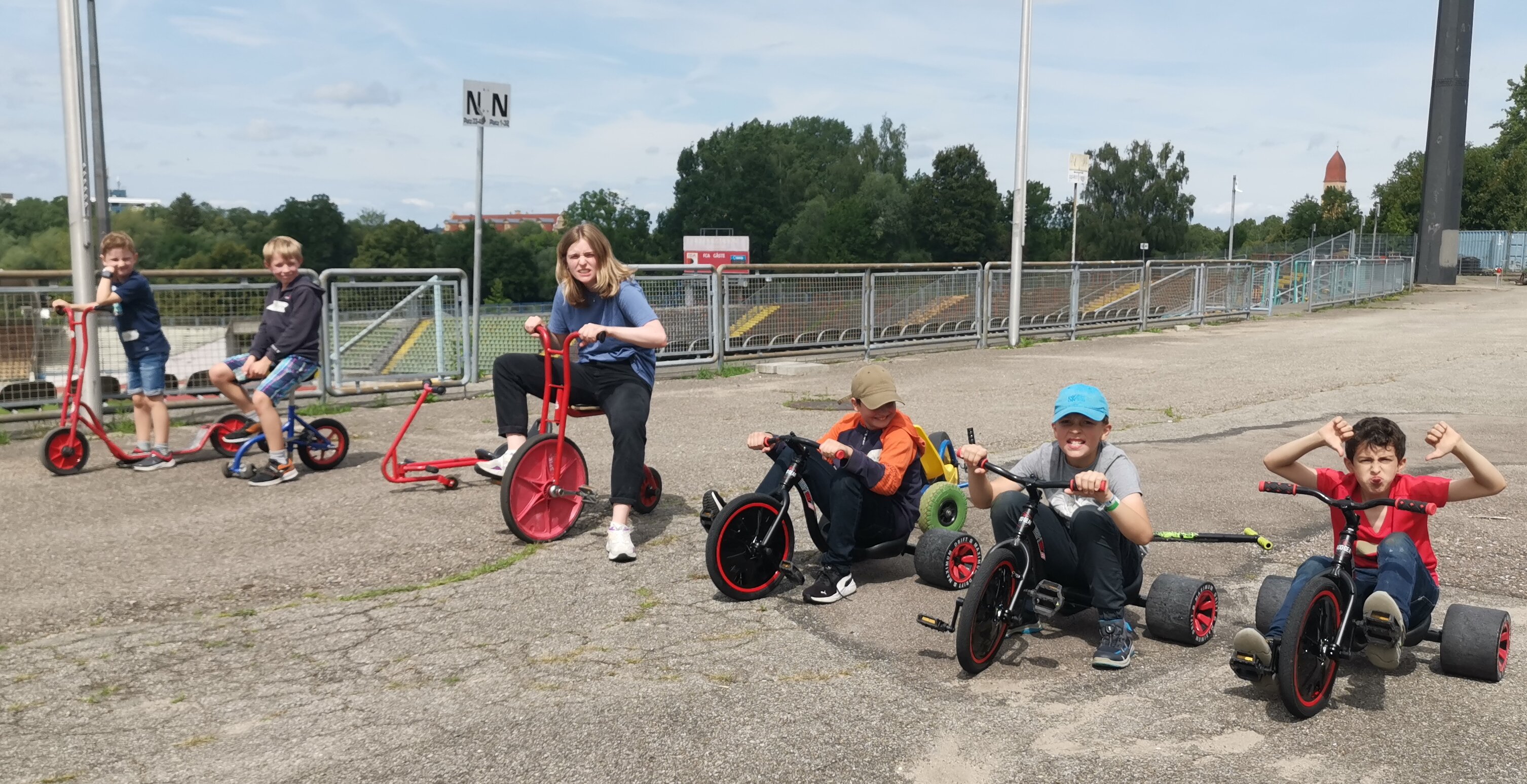 Sechs Kinder sitzen auf lustigen Fahrzeugen wie übergroßem Dreirädern oder Go-Carts. Sie sind in einem Fußballstadion