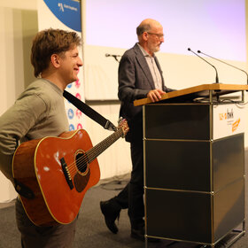 ein Mann mit Glatze und Brille am Rednerpult, daneben ein junger Mann mit Gitarre