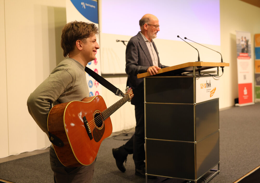 ein Mann mit Glatze und Brille am Rednerpult, daneben ein junger Mann mit Gitarre