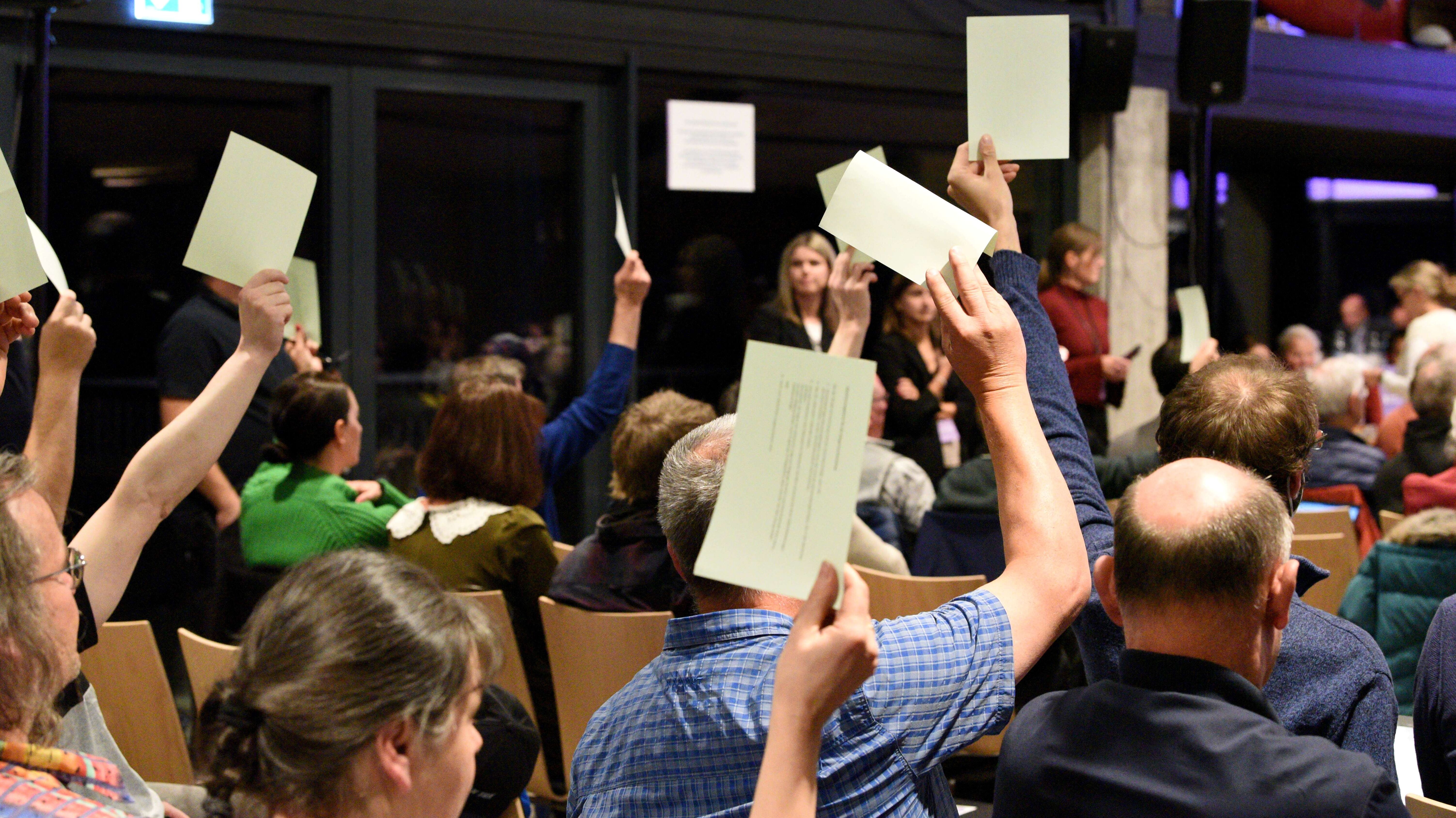 Viele Leute sitzen in einem Saal. Sie heben die Hand zu einer Abstimmung