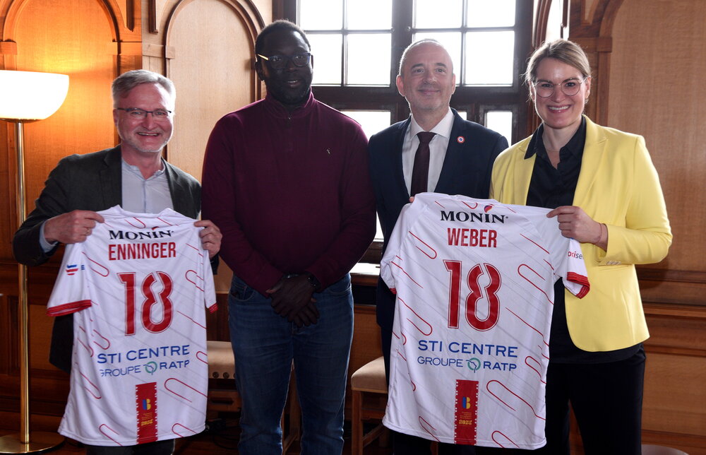 Drei Männer und eine Frau im Rathaus. Ein Mann und die Frau halten Fußballtrikots in der Hand.