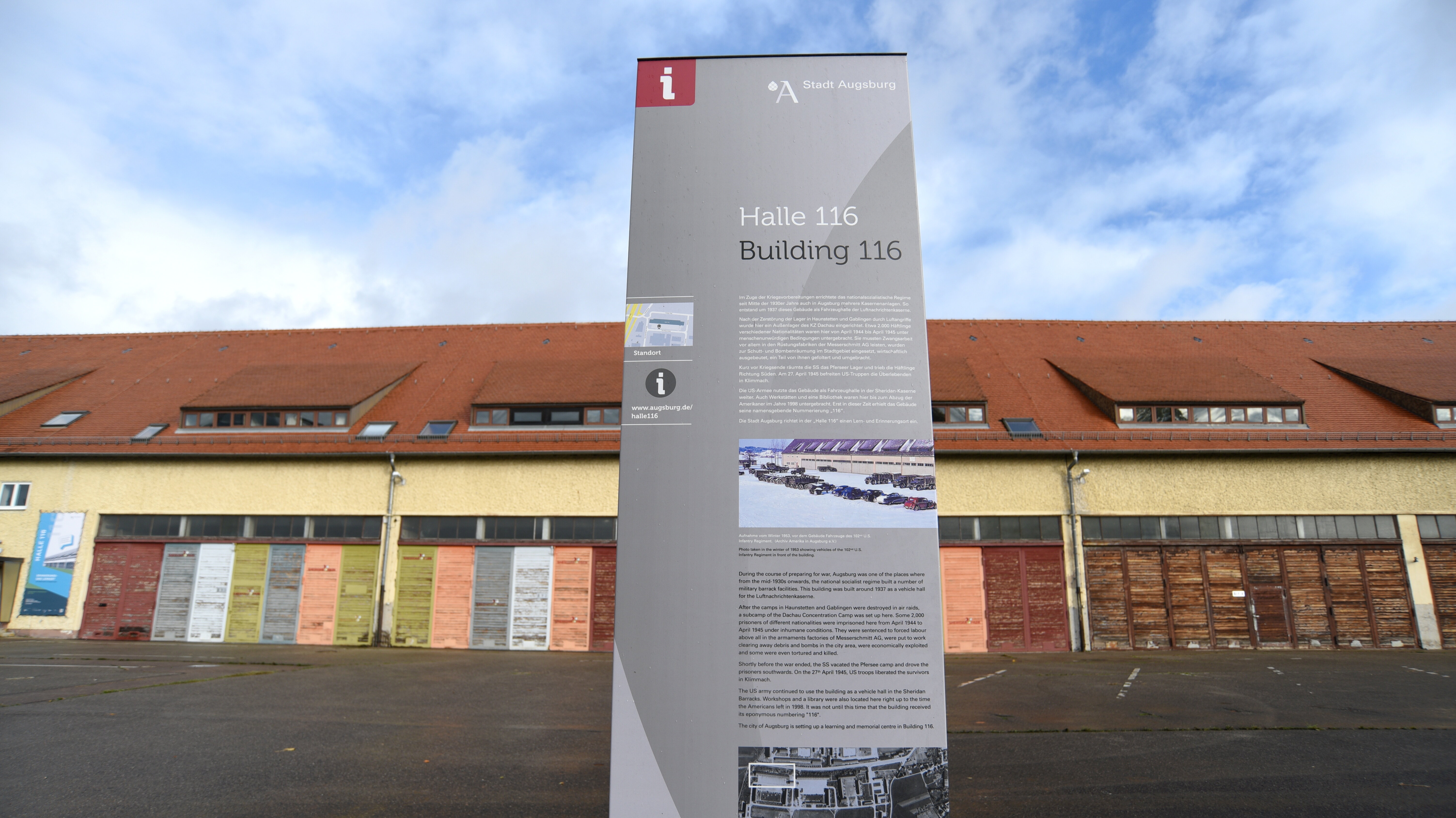 Im Vordergrund steht eine große Informationstafel mit dem Titel Halle 116. Im Hintergrund ist die Halle, ein großes Gebäude mit gelber Fassade und rotem Ziegeldach, zu sehen.