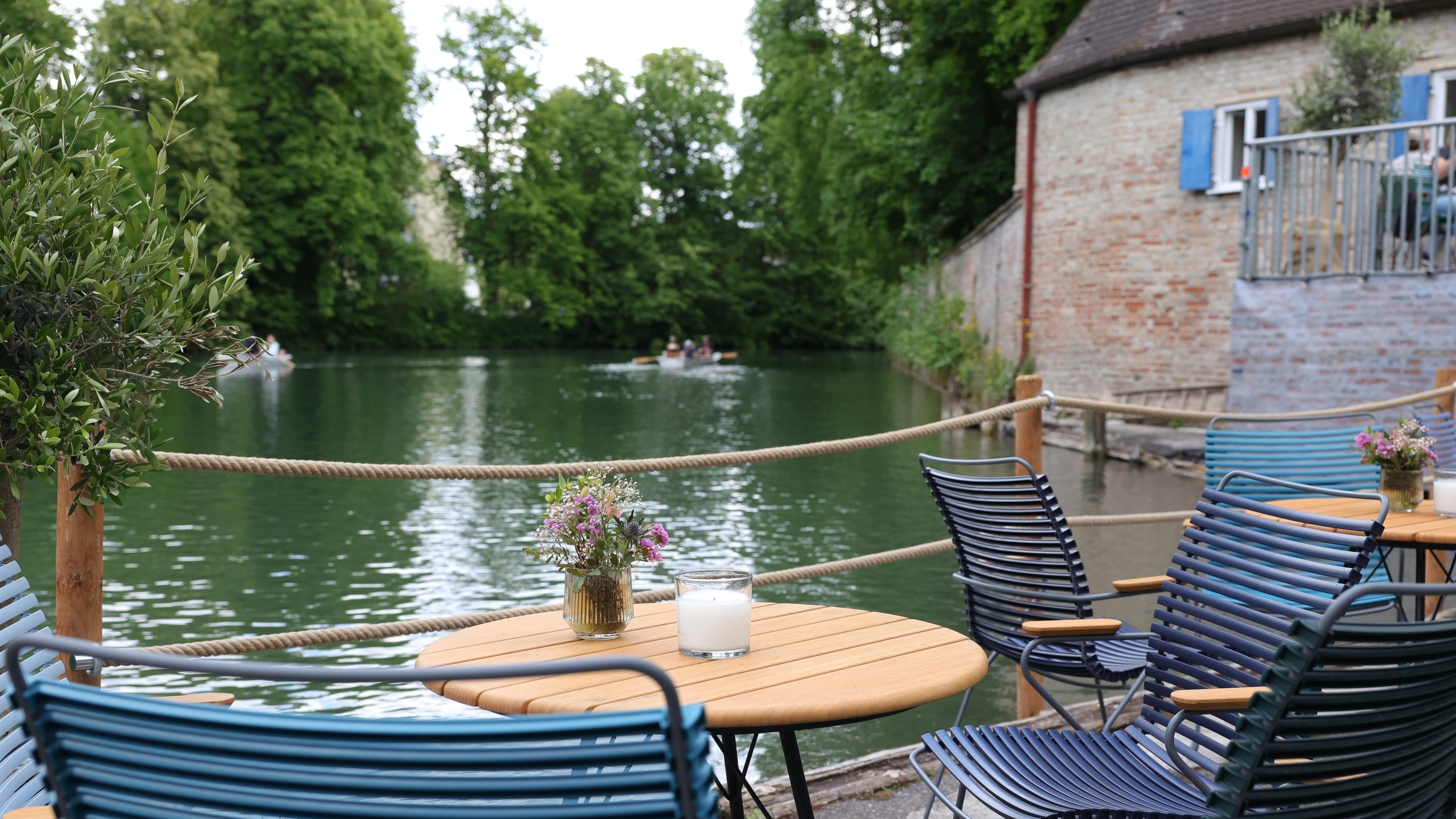 Das Foto zeigt den Biergarten Kahnfahrt. Mehrere dunkelblaue stühle stehen um einen runden Holztisch, auf dem Blumen stehen. Die Stühle stehen auf einer Holzplattform. Von dort hat man Blick auf Wasser, an das im Hintergrund Bäume anschließen