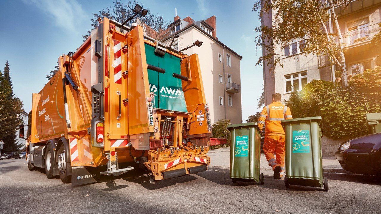 Ein Müllfahrzeug steht vor einem Wohnhaus. Ein Mitarbeiter zieht zwei Mülltonnen zurück in den Hof