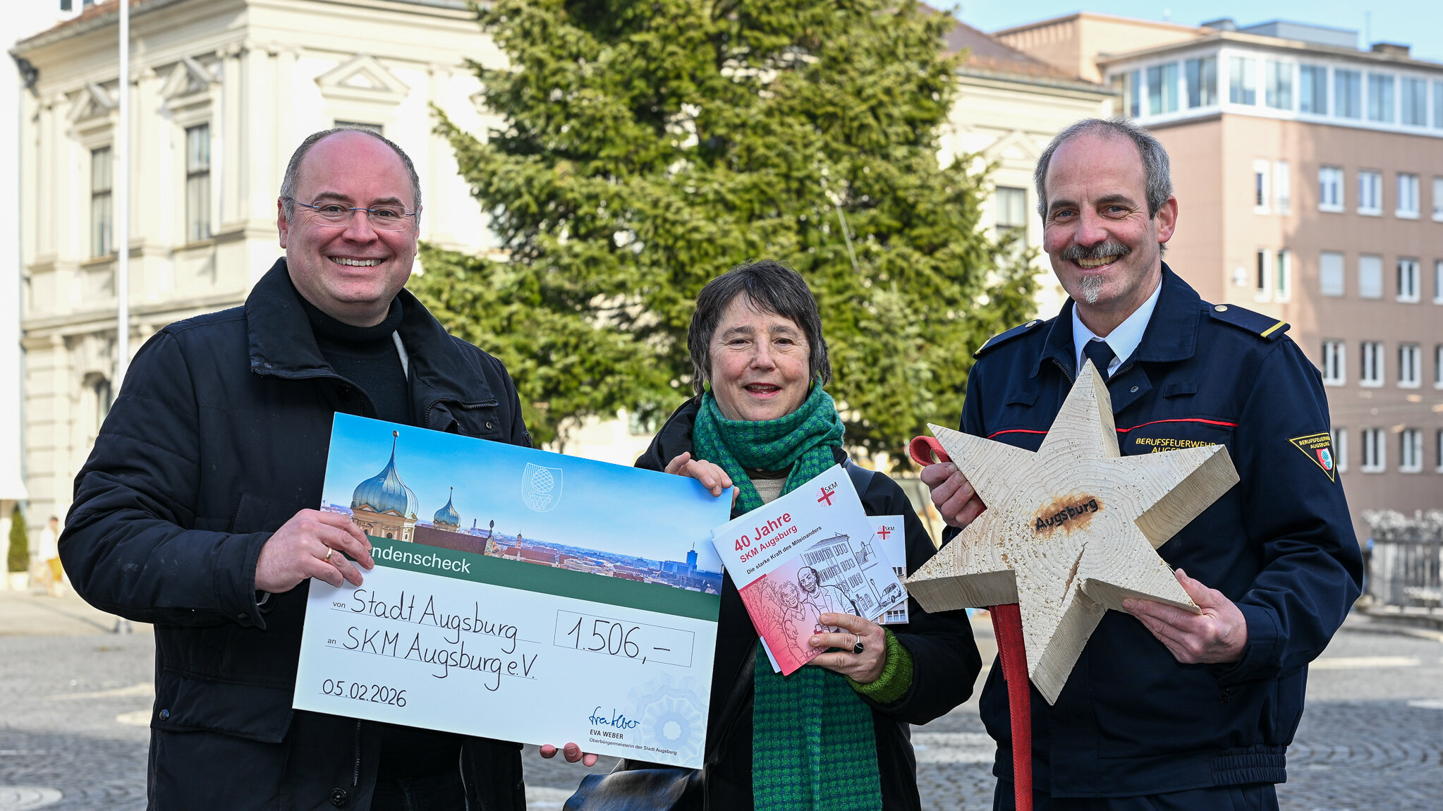 Zwei Männer und eine Frau. Ein Mann gibt ihr einen großen Scheck über 1506 Euro. Der andere Mann hält einen großen Stern aus Holz in der Hand.
