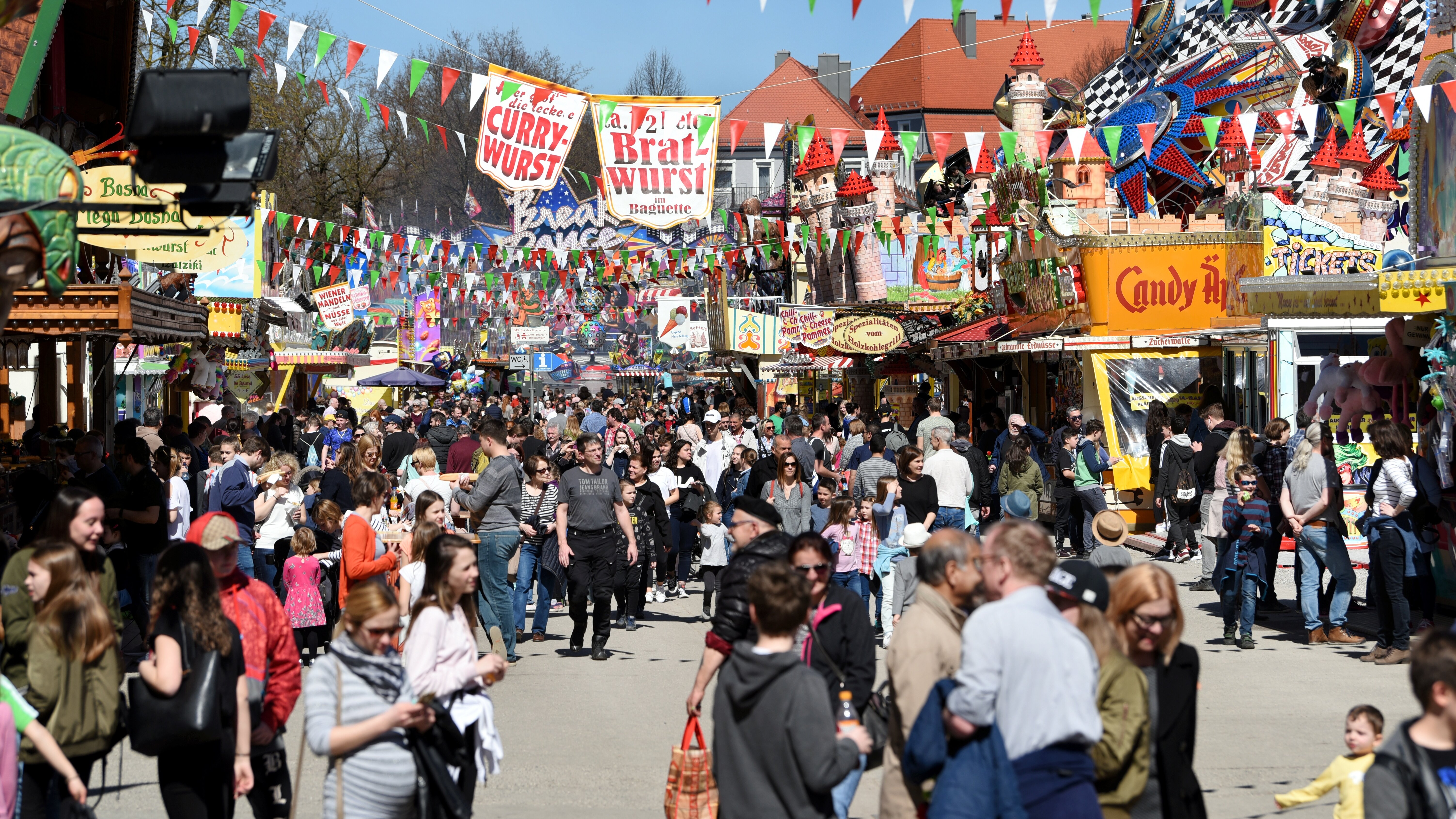 eine Straße gesäumt von Buden und Fahrgeschäften, in der sich viele Leute jeden Alters aufhalten; obendrüber Bänder mit Fähnchen in den Augsburger Stadtfarben