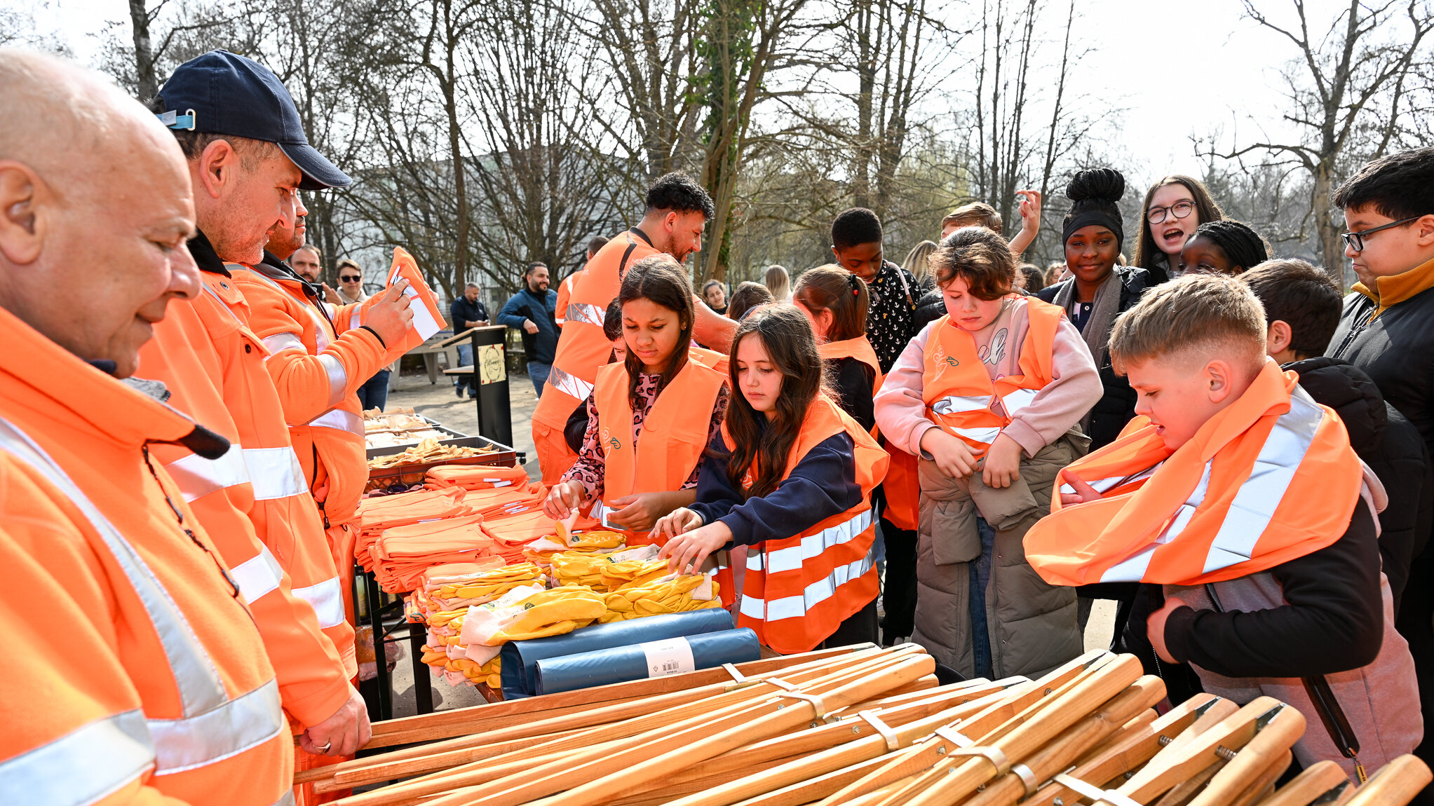 Männer in oranger Arbeitskleidung vor einer Reihe Abfallzangen und Warnwestenauf einem Tisch.  Dahinter Kinder unterschiedlichen Alters, die gerade die Warnwesten anziehen.