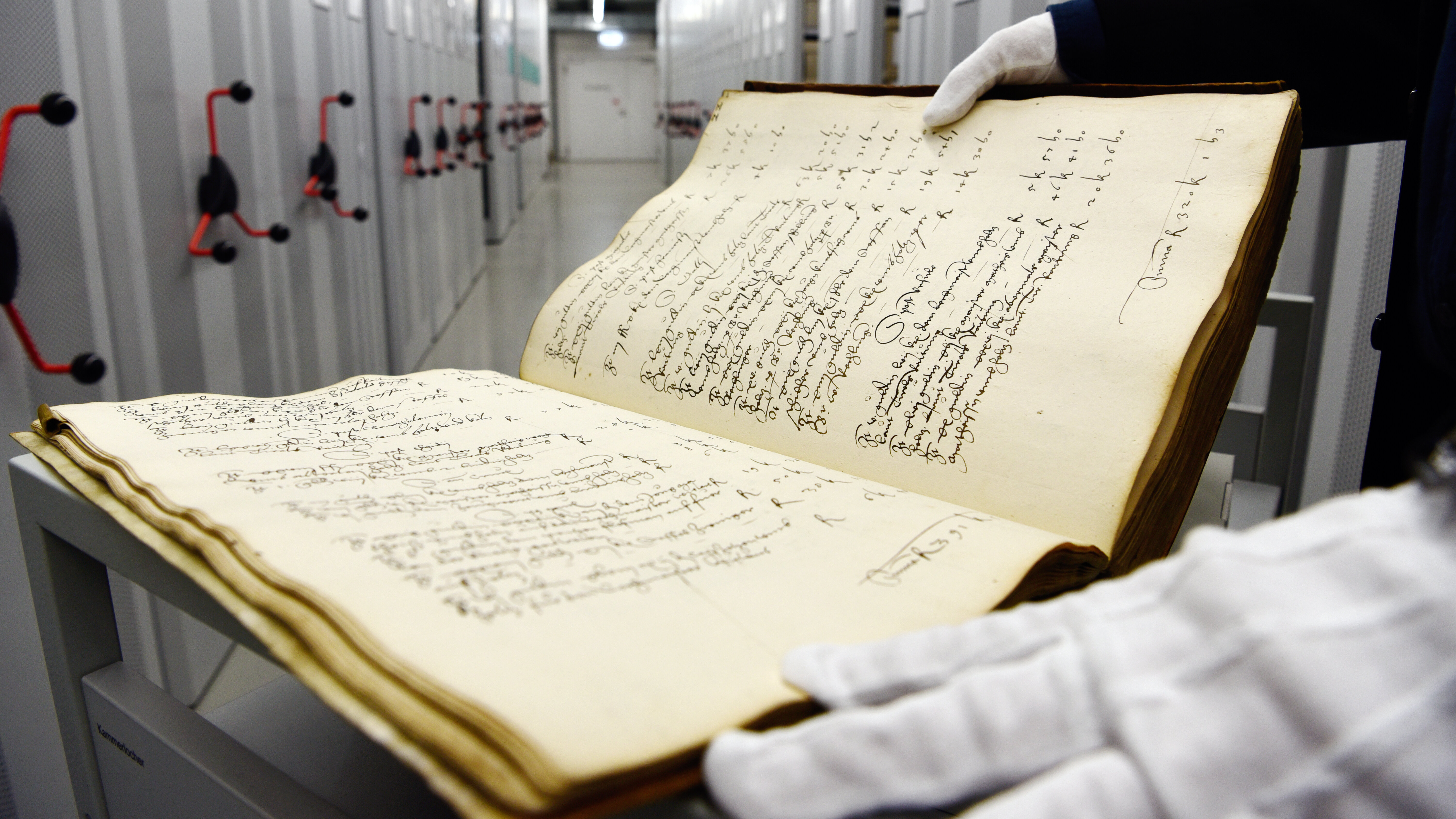Das Foto zeigt ein sehr altes Buch mit schnörkeliger Schrift und vergilbten Seiten. Im Hintergrund sind Regale des Stadtarchivs zu sehen.