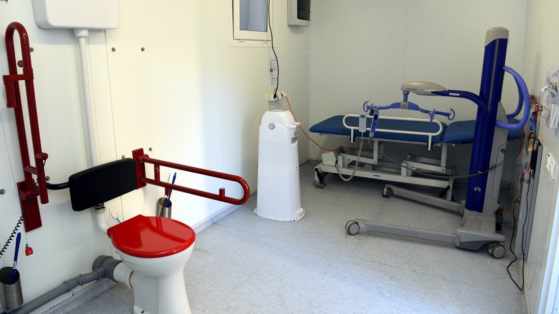 Ausstattung eines „Toilette für Alle“-Containers von Innen. Mit Toilettenschüssel mit Stützen auf jeder Seite, einer höhenverstellbaren Pflegeliege und einem Personenstandlifter sowie ein Mülleimer.