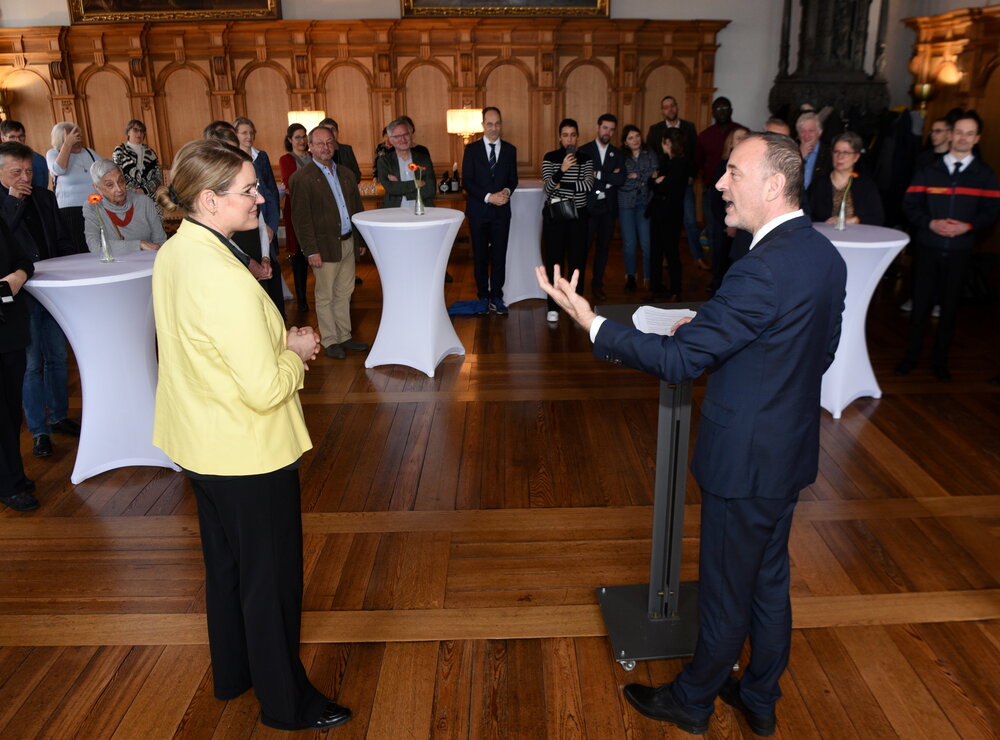 Eine Frau und ein Mann im Rathaus, im Hintergrund rund 20 weitere Personen