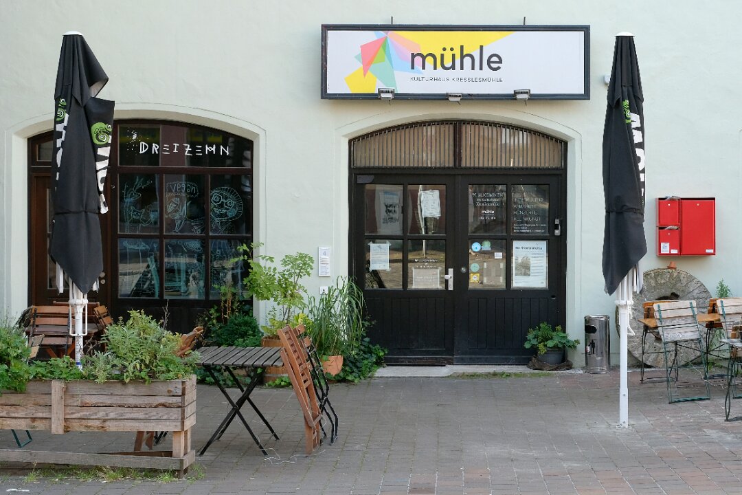 Frontalfoto des Eingangs der Kresslesmühle. Über der großen hölzernen Eingangstüre hängt ein Schild mit dem Namen. Links und rechts sieht man Tische, Stühle und Schirme des Restaurants dreizehn. 