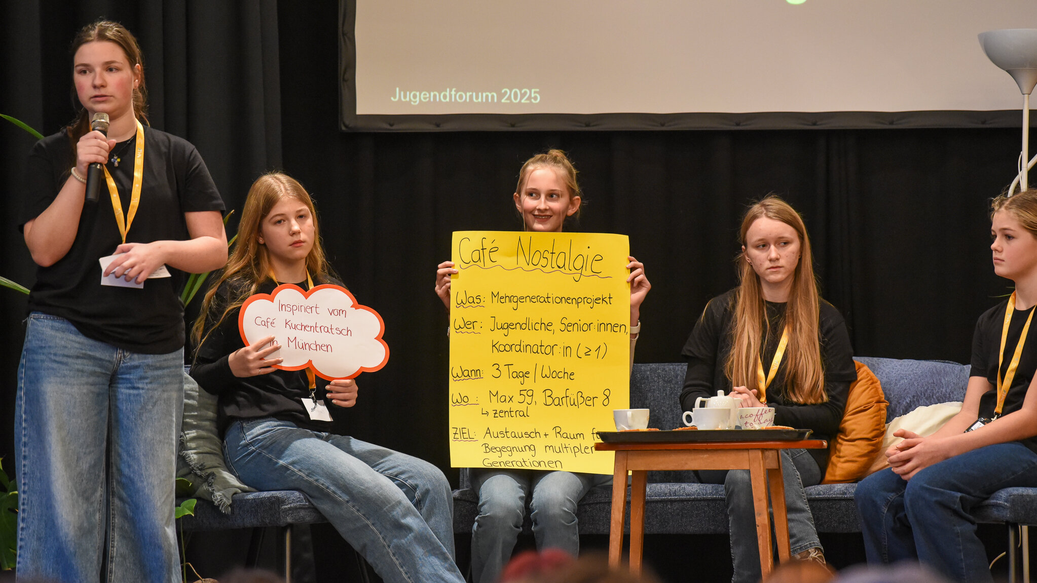 Fünf Mädchen sind auf einer Bühne. Vier von ihnen sitzen auf einem Sofa. Ein Mädchen hält ein gelbes Plakat mir der Überschrift Café Nostalgie. 