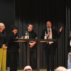drei Frauen und drei Mäner um einen Stehtisch; einer der Männer hält ein Mikrofon in der Hand und spricht, die andere Hand hat er erhoben