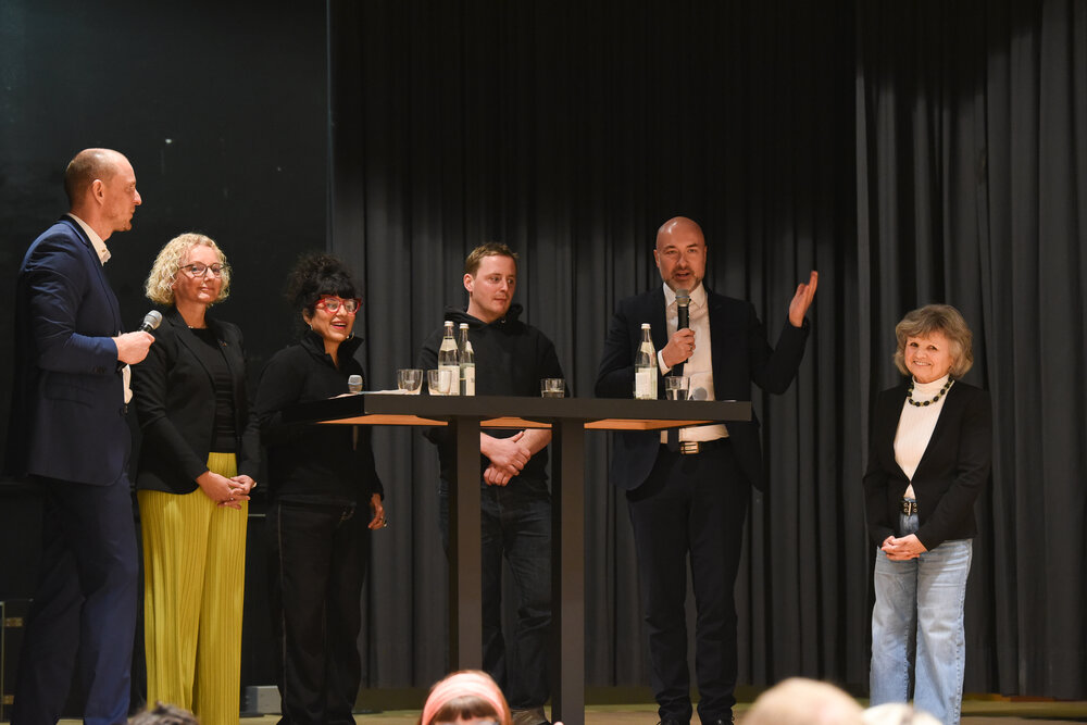 drei Frauen und drei Mäner um einen Stehtisch; einer der Männer hält ein Mikrofon in der Hand und spricht, die andere Hand hat er erhoben