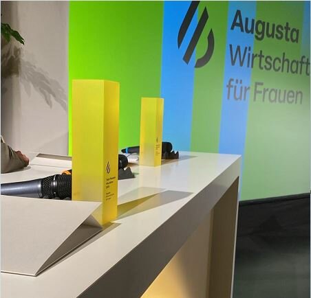 ein Tisch, auf dem zwei gelbe Säulen im quadratischen Grundriss stehen, dahinter ein Plakat mit der Aufschrift Augusta - Wirtschaftspreis für Frauen