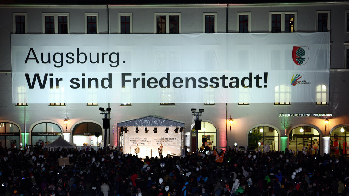 viele Menschen in der Dunkelheit auf einem Platz; am angrenzenden Gebäude ein riesiges, hell angetrahltes Transparent mit der Aufschrift "Augsburg. Wir sind Friedensstadt!"