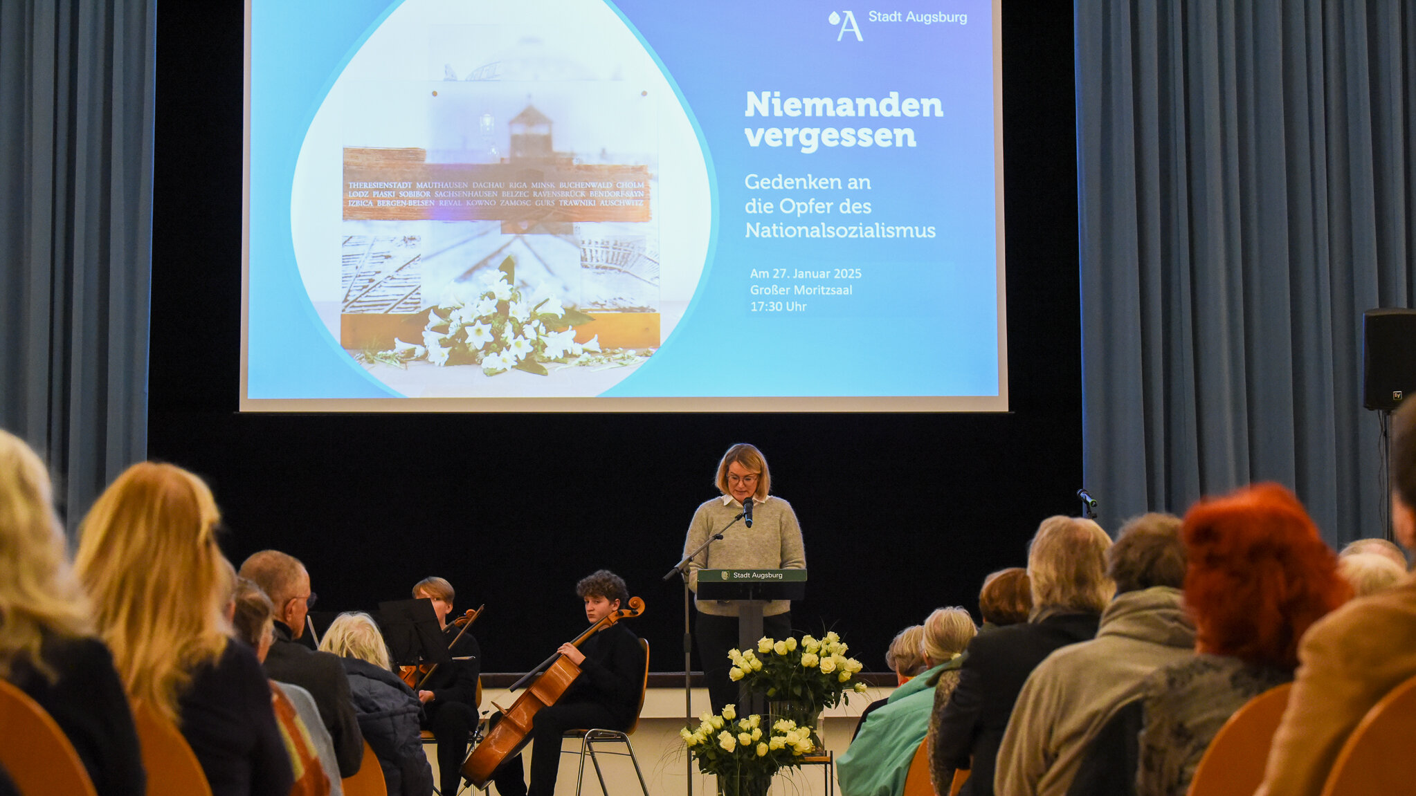 blonde Frau steht am Rednerpult auf einer Bühne, hinter ihr auf der Leinwand die Worte: Niemanden vergessen